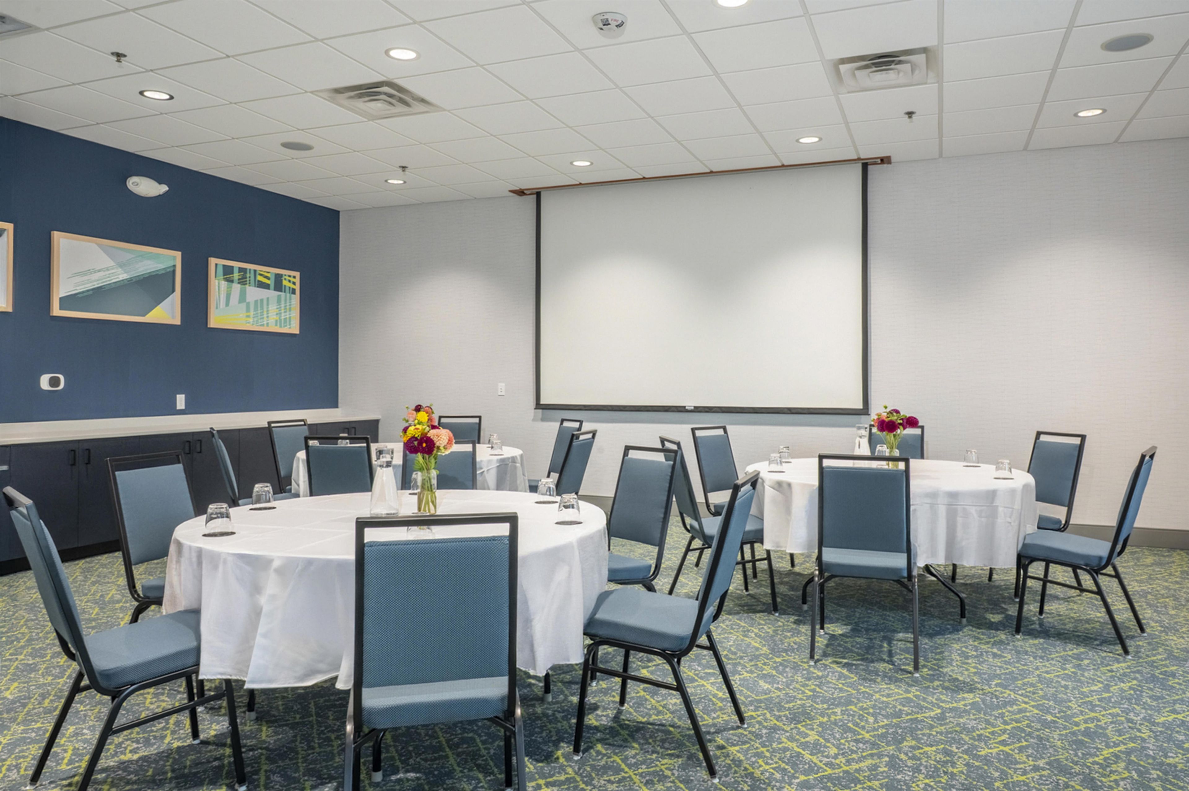 A conference room with round tables, chairs, a projector screen, and two framed artworks on the wall.