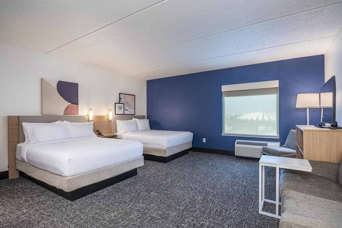 A hotel room with two beds, a desk, a lamp, and a large window.