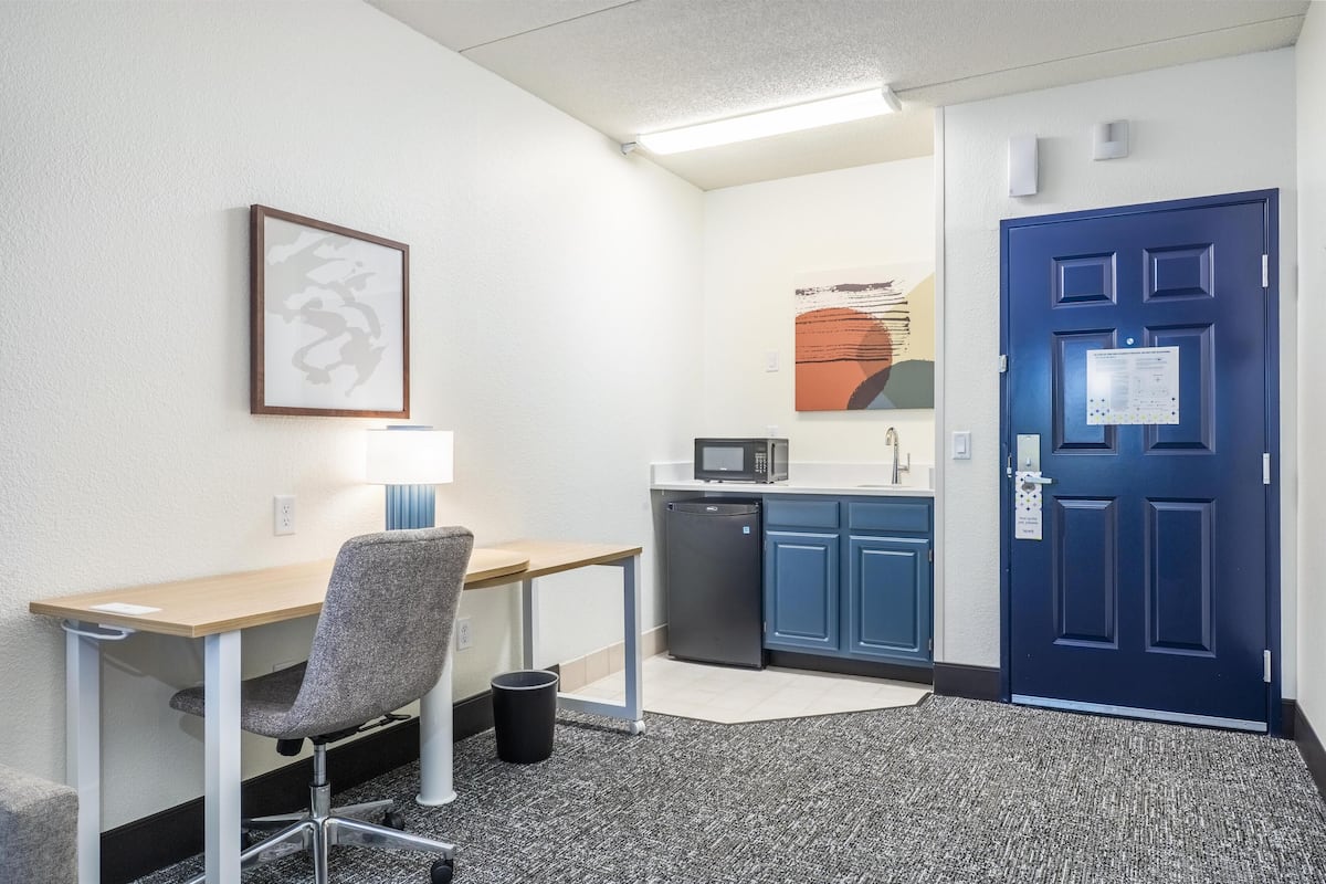 A small kitchenette with a microwave, refrigerator, and sink. There is a desk with a chair and a trash can. A blue door is visible in the background.