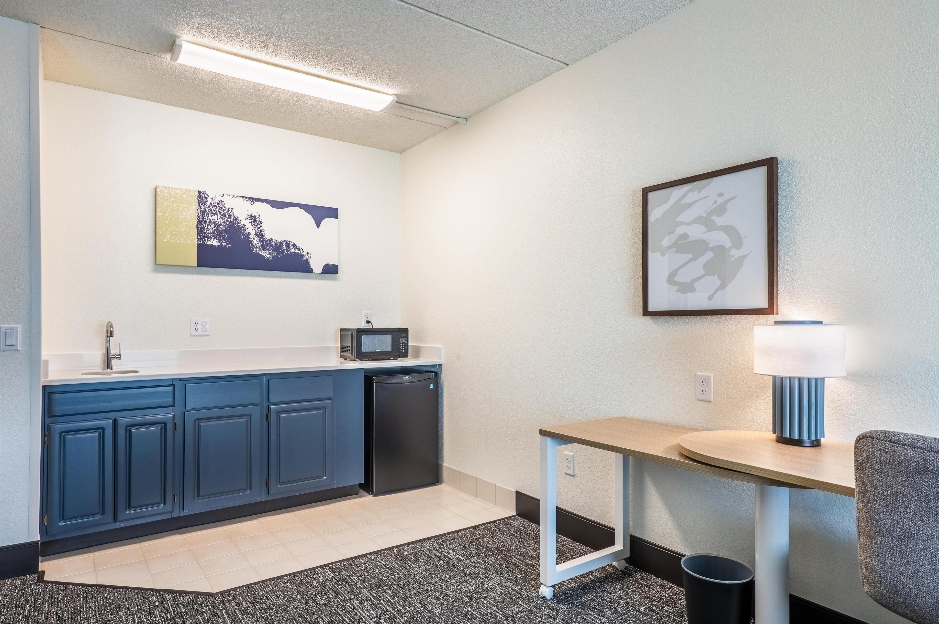 A compact kitchenette with a microwave, sink, and refrigerator, a small table with a lamp, and a framed picture on the wall.