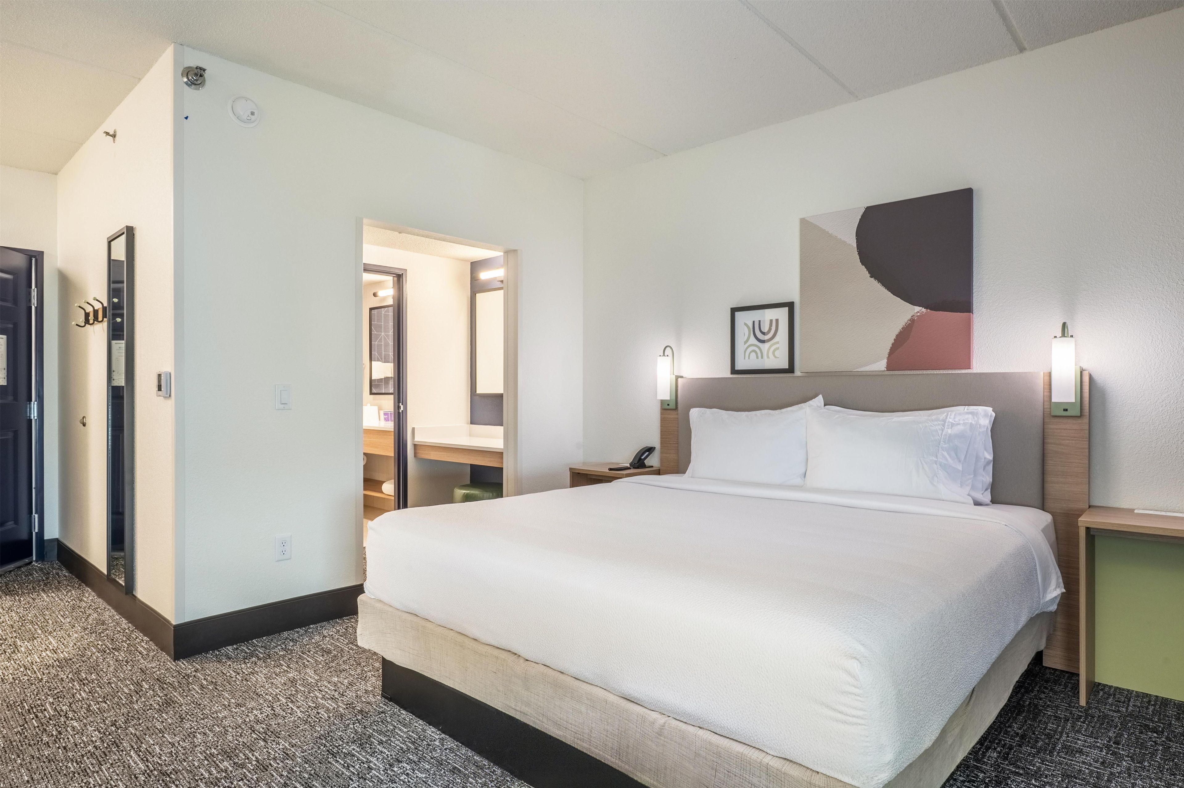 Una habitación de hotel con una cama, una mesita de noche, un teléfono en la pared, un espejo y una puerta que da a un baño.