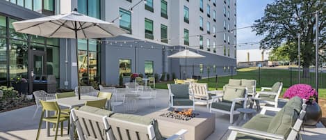 An outdoor seating area with tables, chairs, and umbrellas, surrounded by a modern building with large windows.