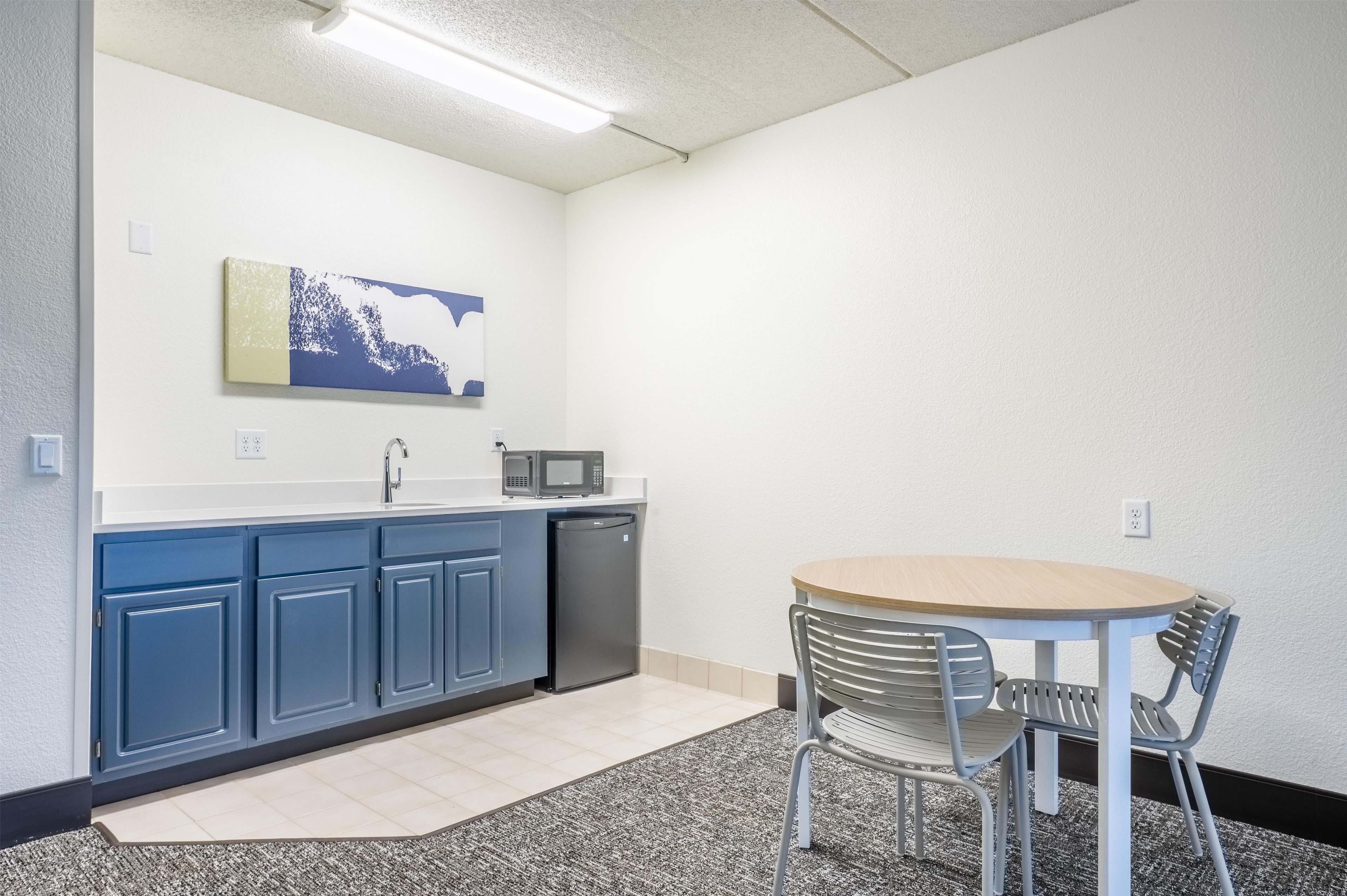 Una pequeña kitchenette con microondas, pileta y armarios. Hay una mesa redonda con dos sillas y una alfombra en el suelo.