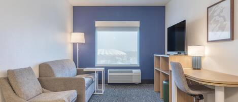 A hotel room with a sofa, a desk with a chair, a TV, a lamp, and a window with a view of a building.