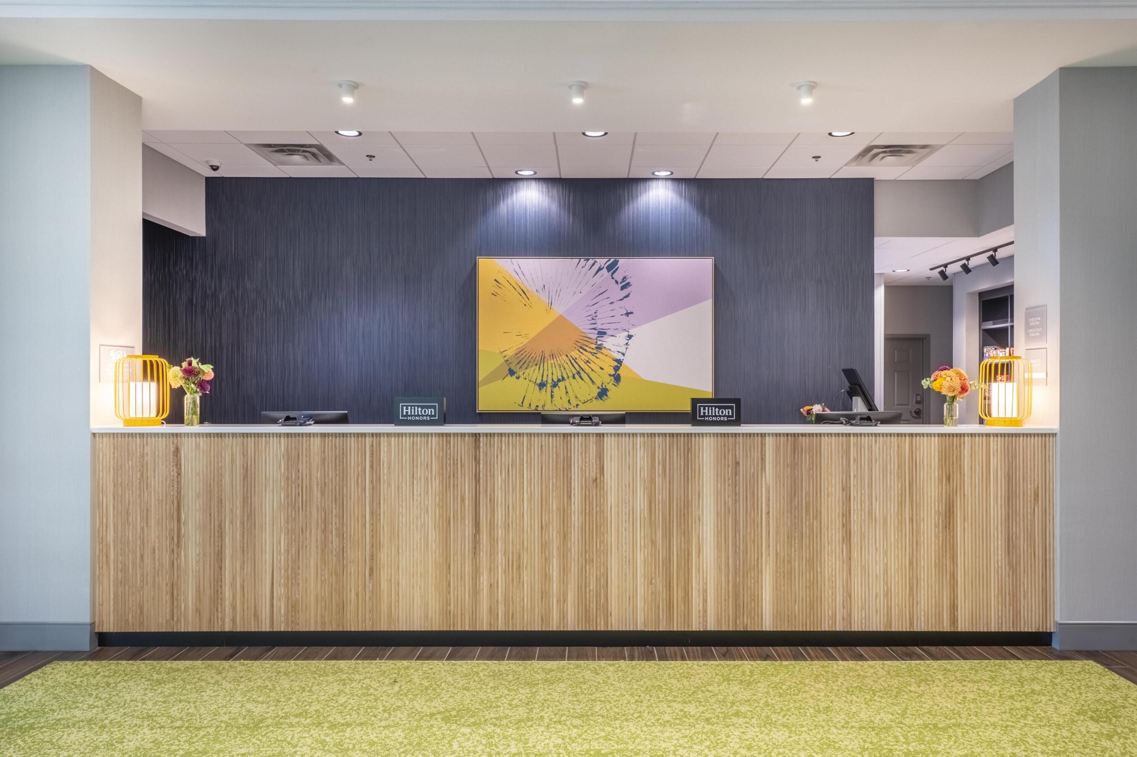 A hotel reception desk with a modern design, featuring a wooden panel front, a large abstract artwork above the counter, and two illuminated lamps on either side.