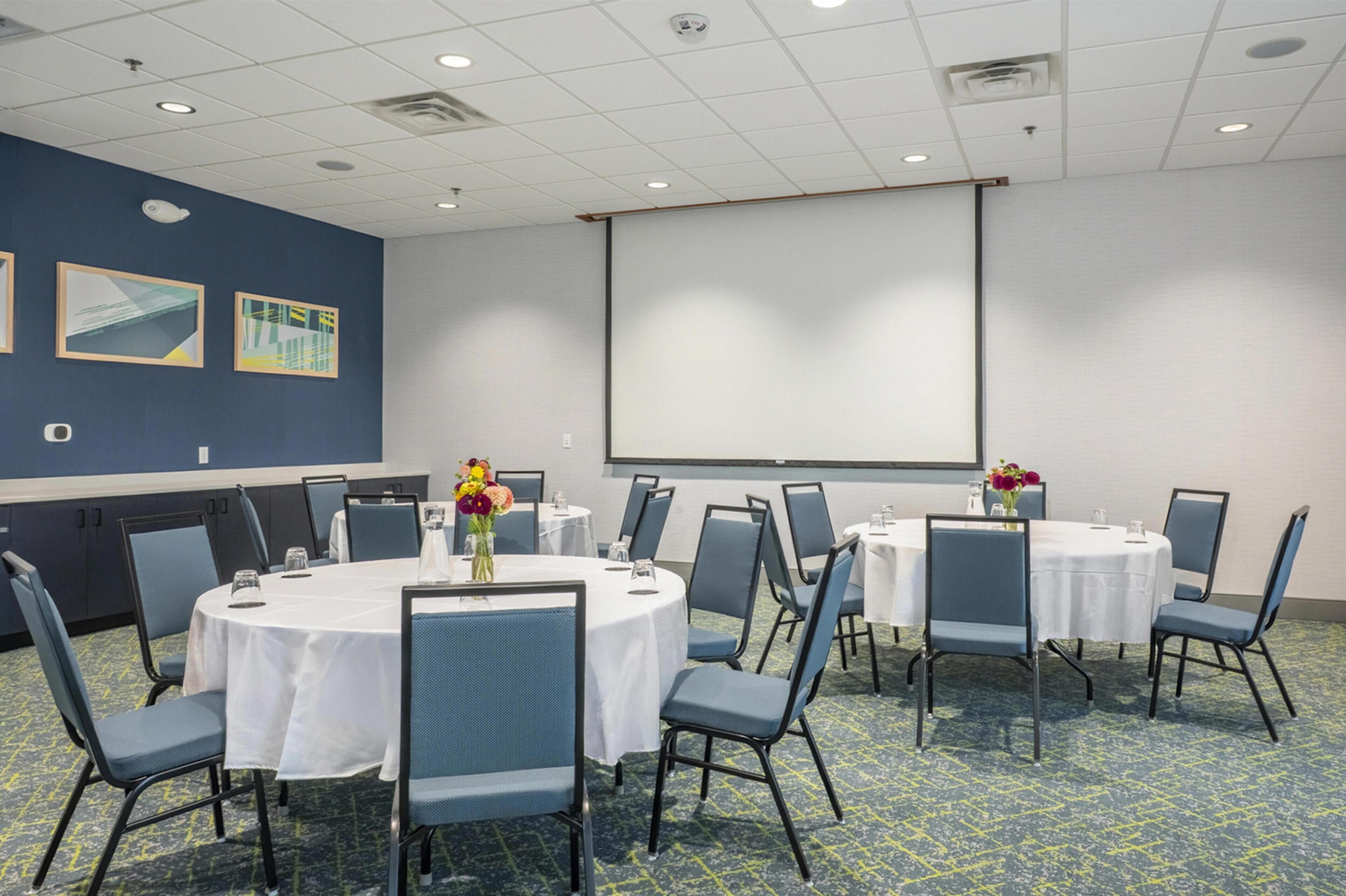 A conference room with round tables, blue chairs, and a projector screen on the wall.