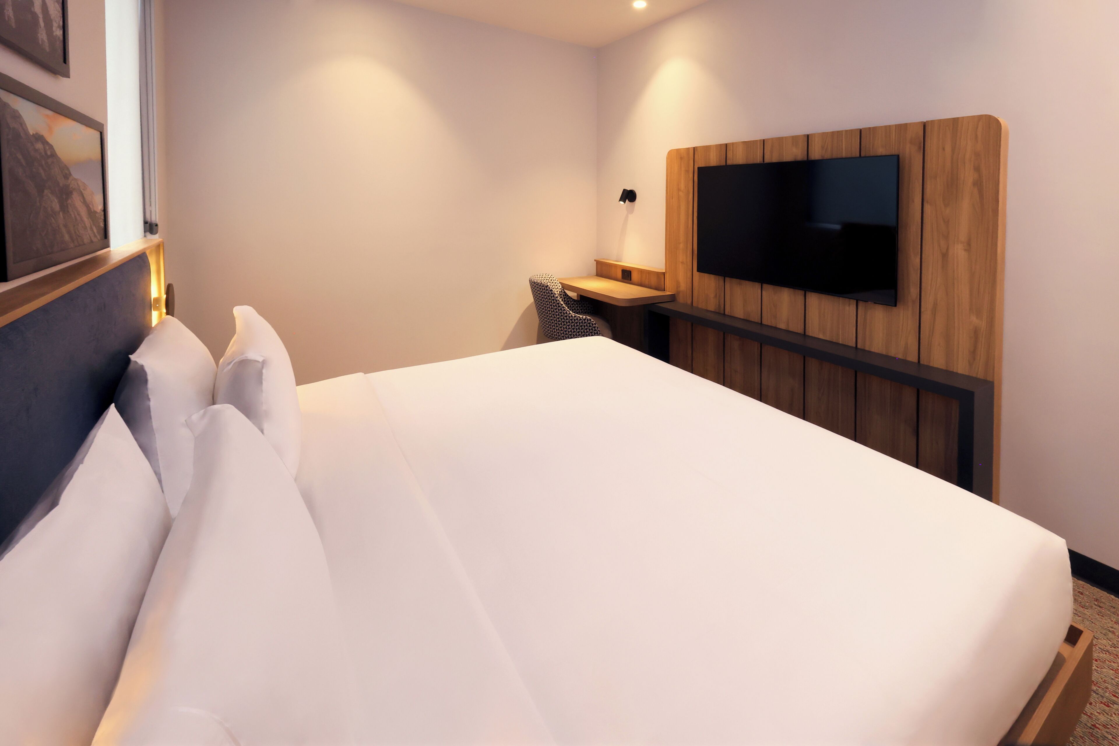 A modern hotel room with a neatly made bed, a flat-screen TV mounted on a wooden panel, and a small desk with a chair.