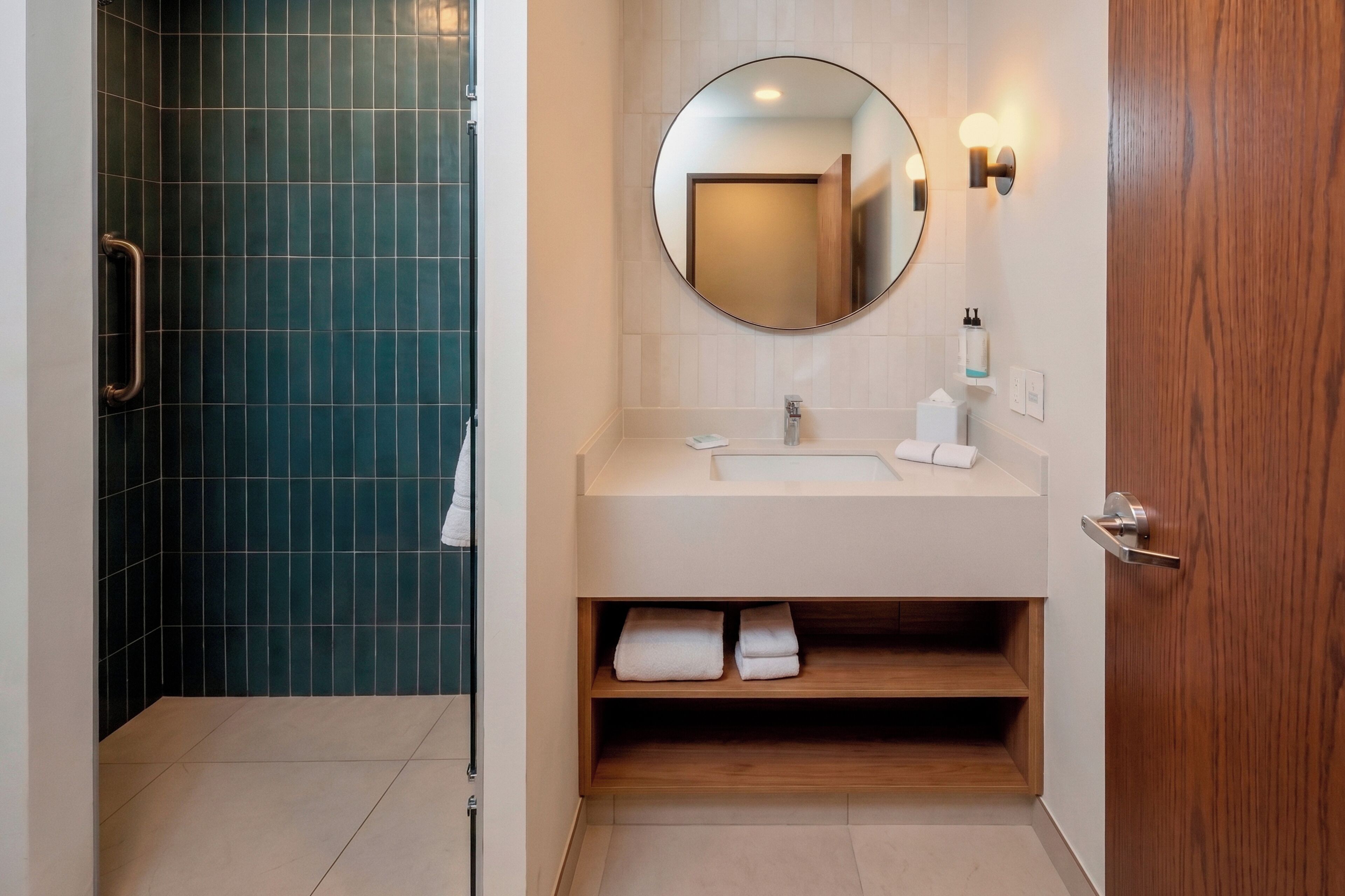 A bathroom with a shower, a sink, a mirror, and a wooden shelf.