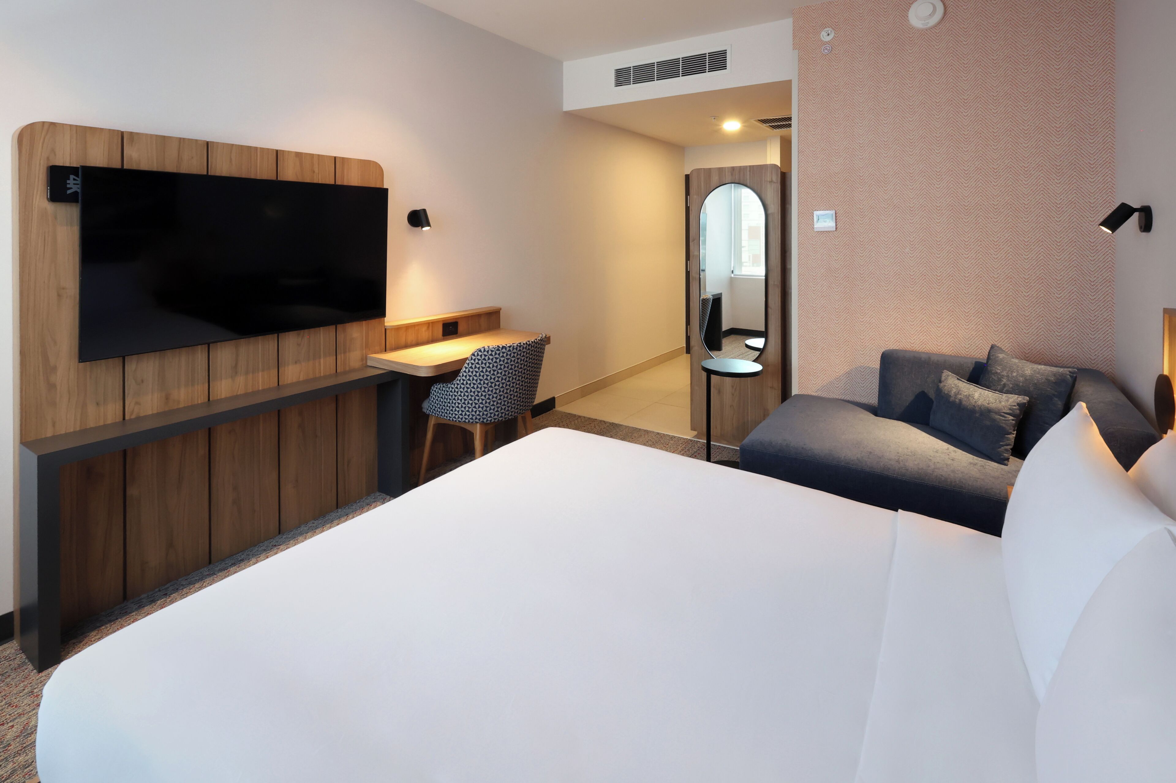 Hotellihuone, jossa on sänky, työpöytä tuolilla, seinälle asennettu televisio, peili ja sohva.
