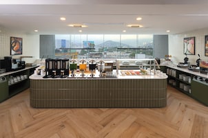 Un café moderno con un mostrador central, varias máquinas de café y vistas de la ciudad a través de amplias ventanas.