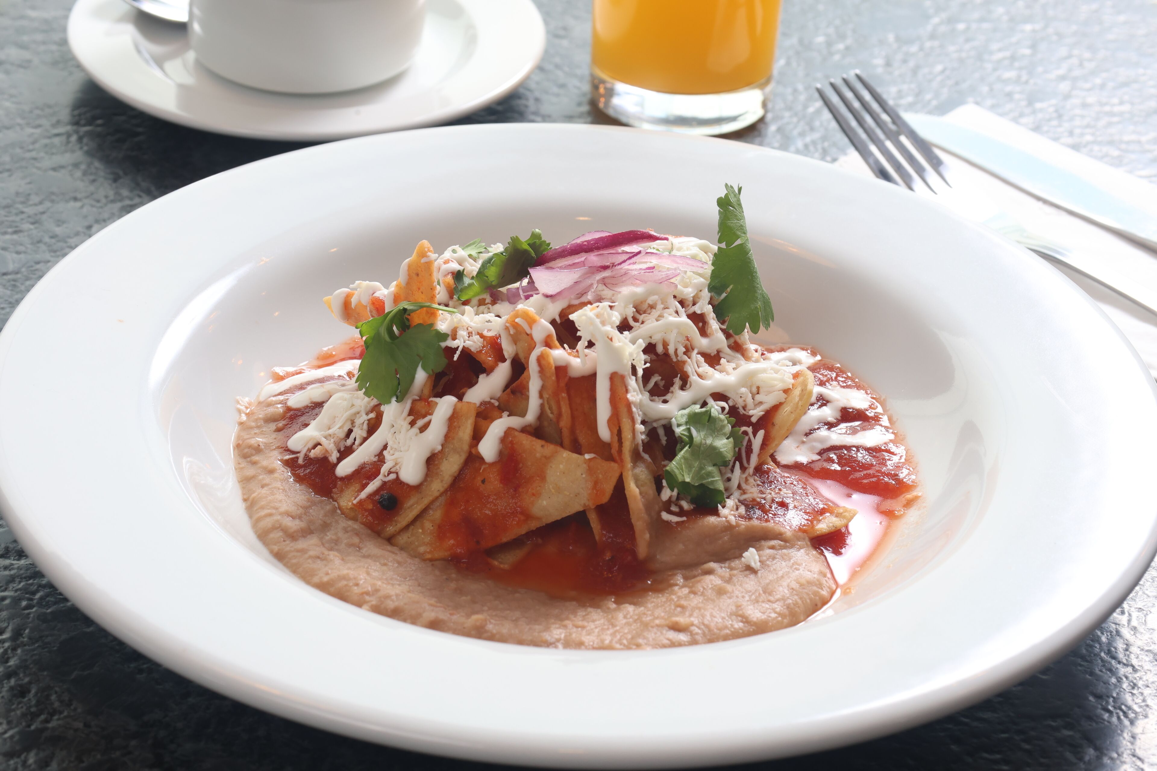 Un plato de enchiladas con ingredientes y salsa, una taza y un vaso de jugo de naranja sobre una mesa.