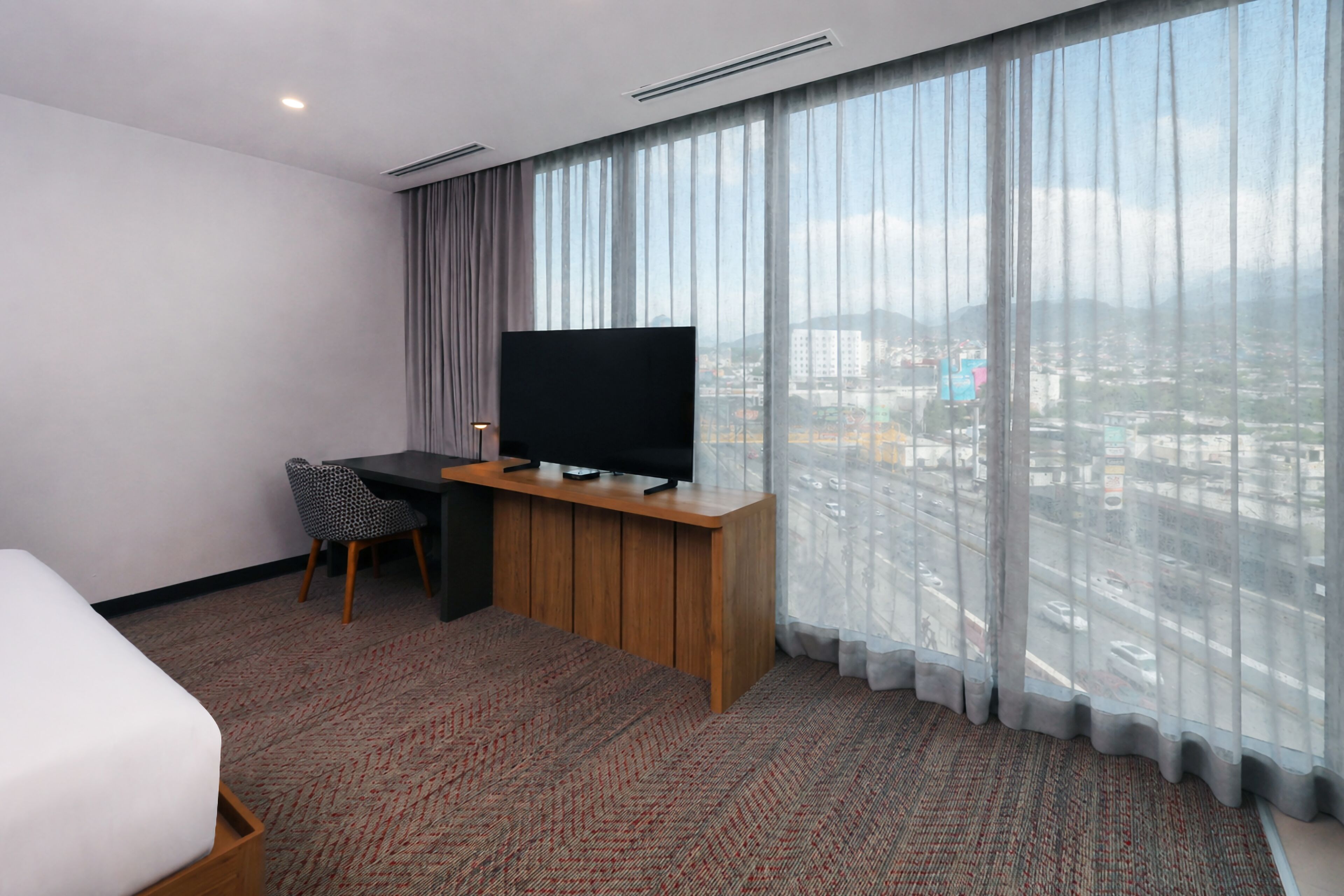 A hotel room with a bed, a desk with a television, a chair, and a large window with a city view.