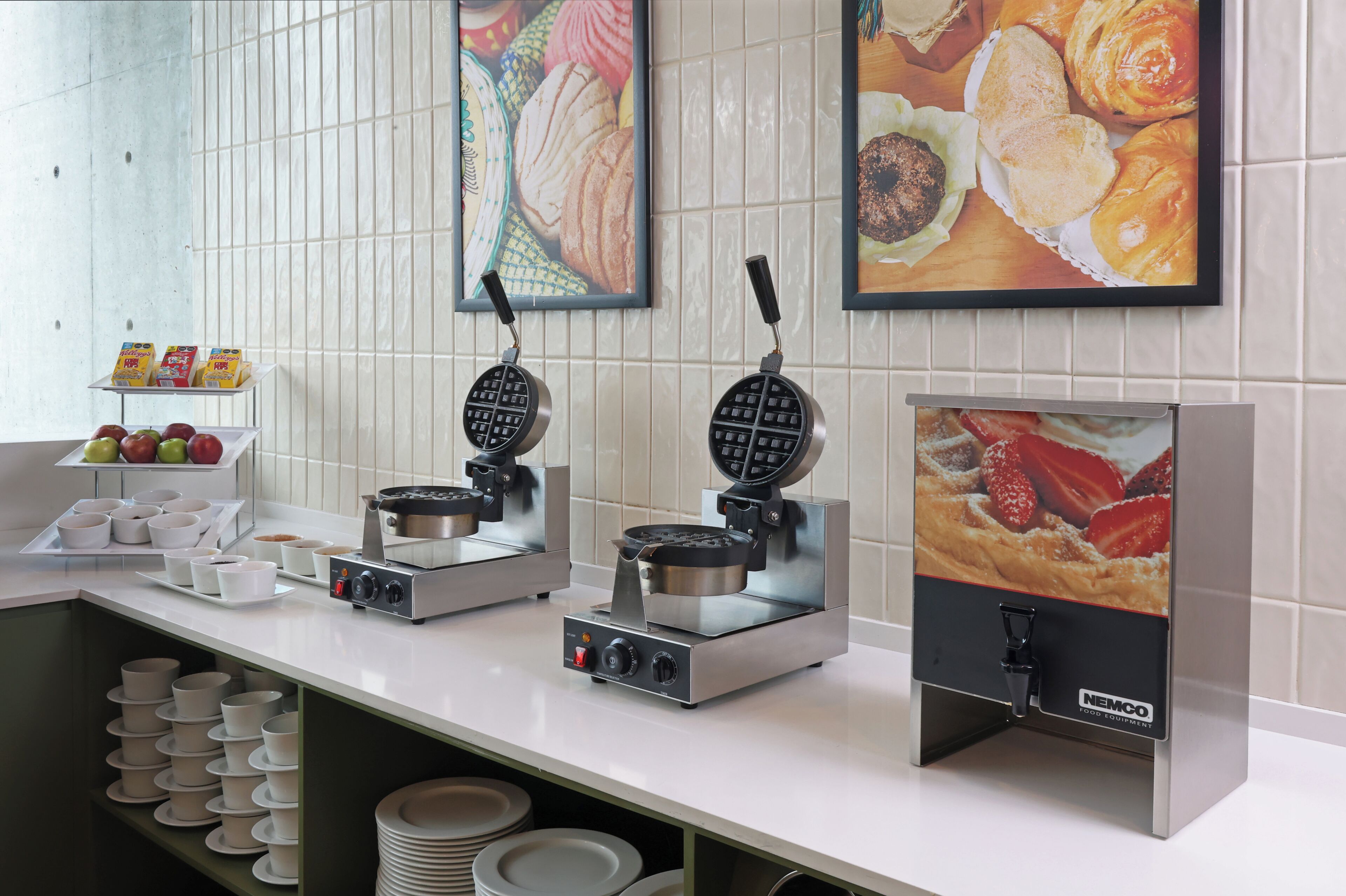 A breakfast buffet setup with waffle makers, a fruit display, and a wall-mounted food display.