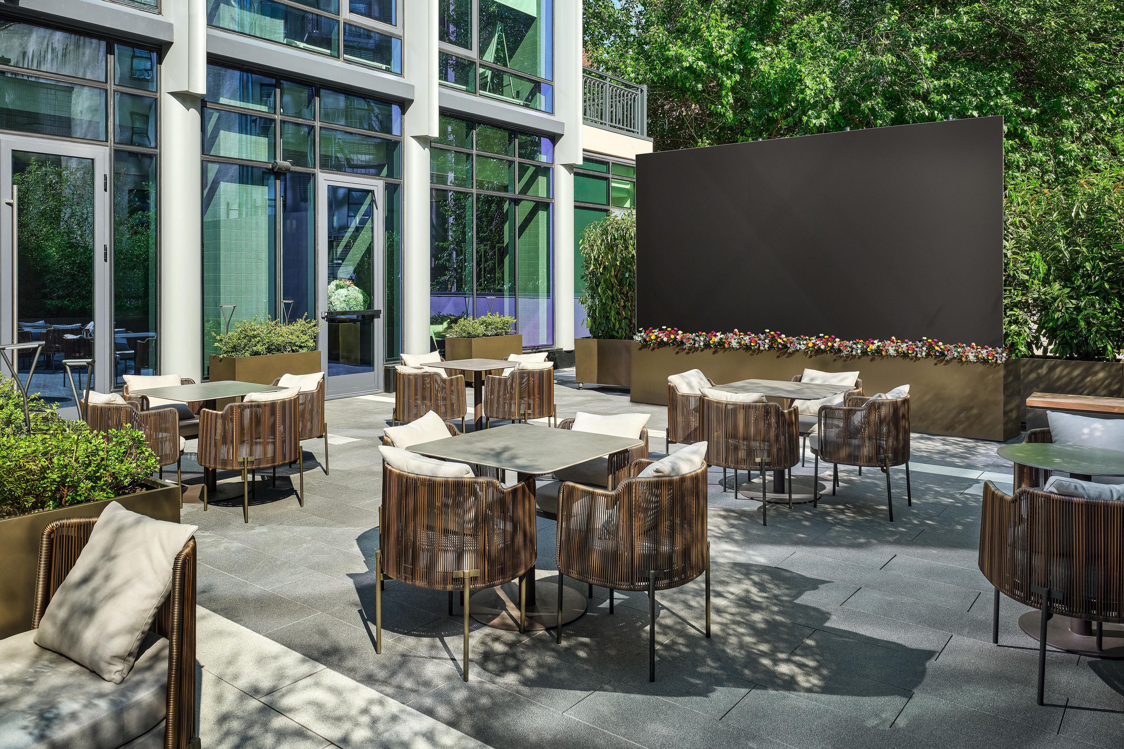 An outdoor seating area with tables and chairs, surrounded by greenery and a building with large windows.