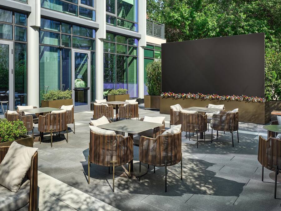 An outdoor seating area with tables and chairs, surrounded by greenery and a building with large windows.