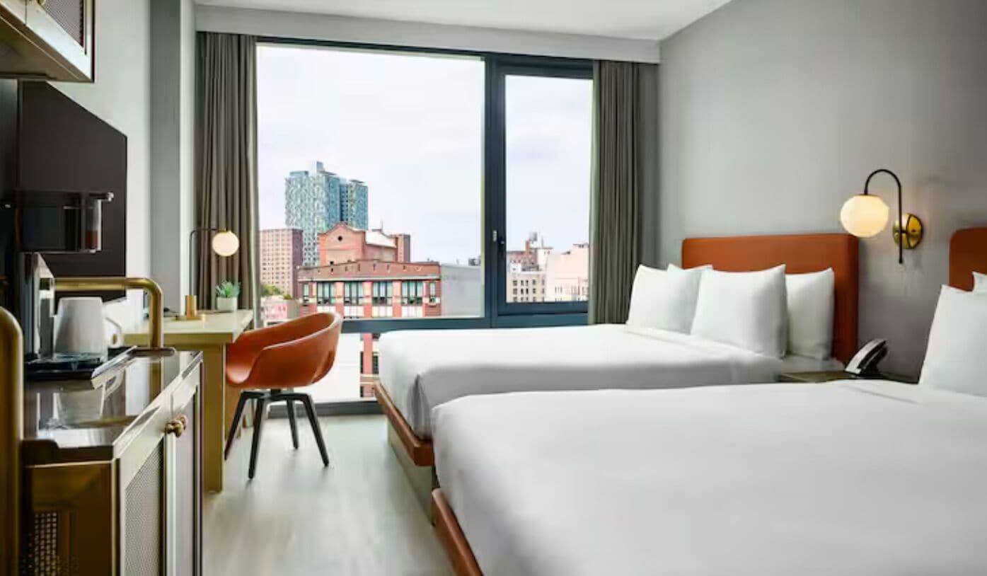 A hotel room with two beds, a desk with a chair, a window with a city view, and a wall-mounted lamp.