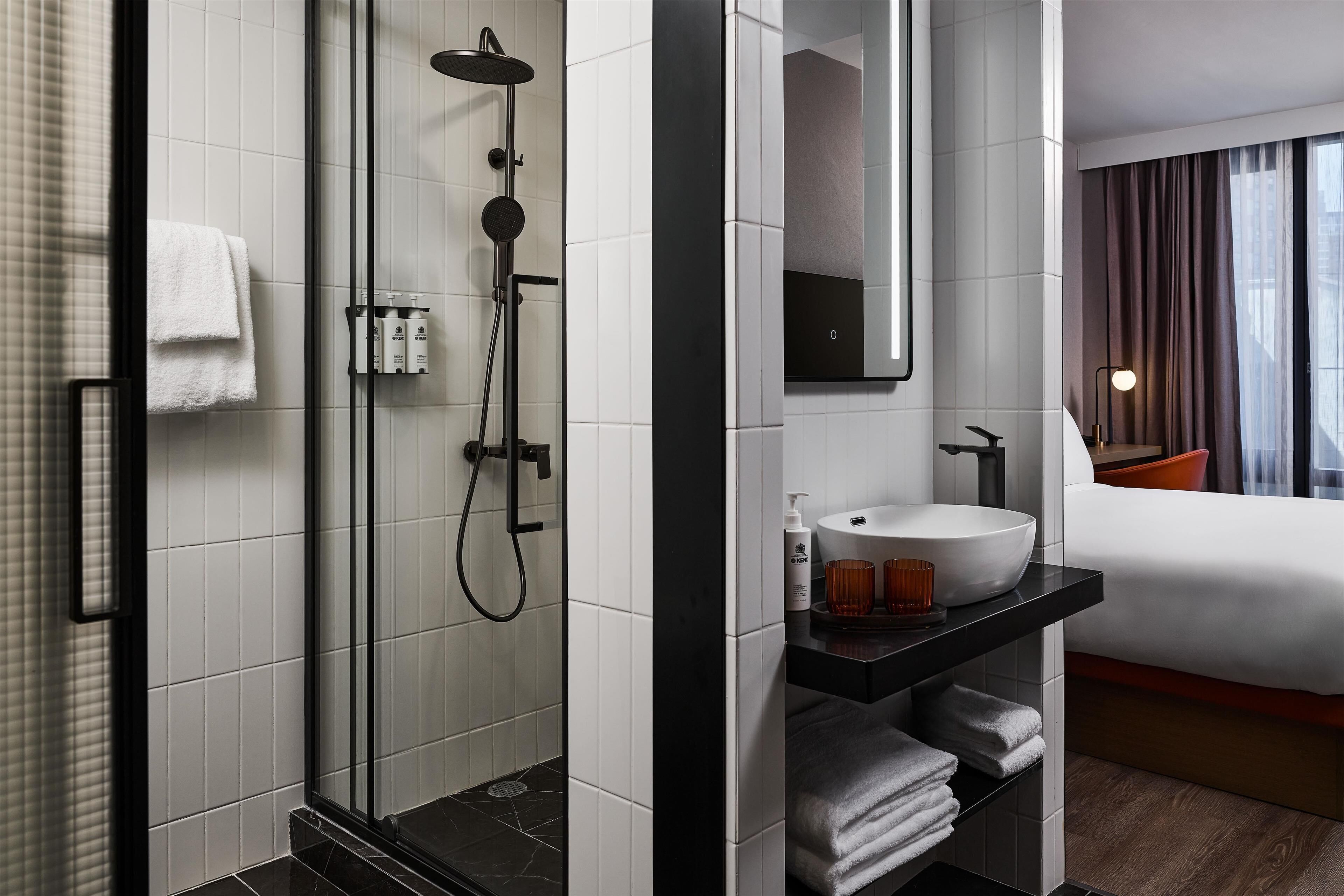 A modern hotel bathroom with a glass-enclosed shower, a white sink, and a bed with a view of the city.