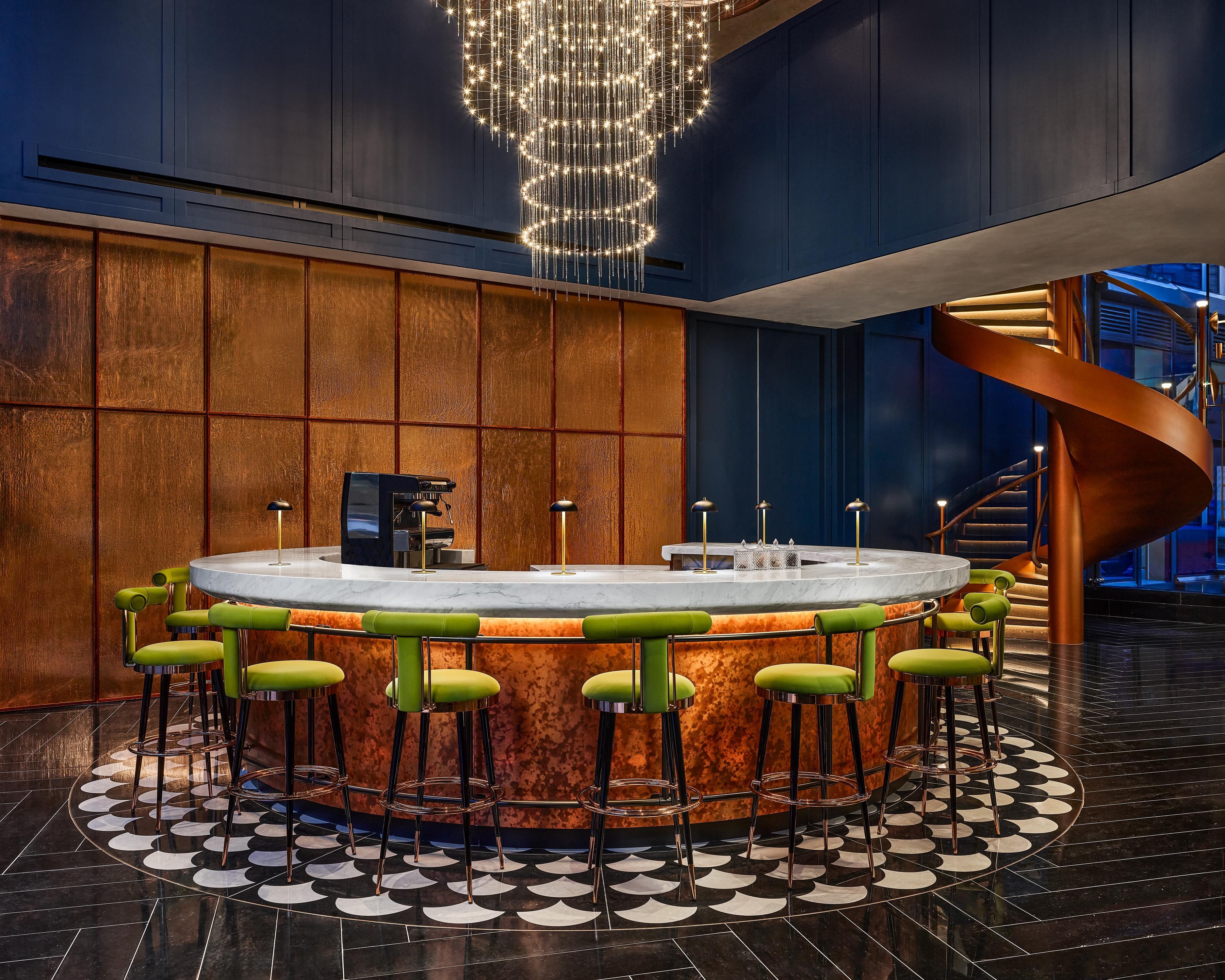 A bar area with a circular bar, green bar stools, a chandelier, and a spiral staircase.