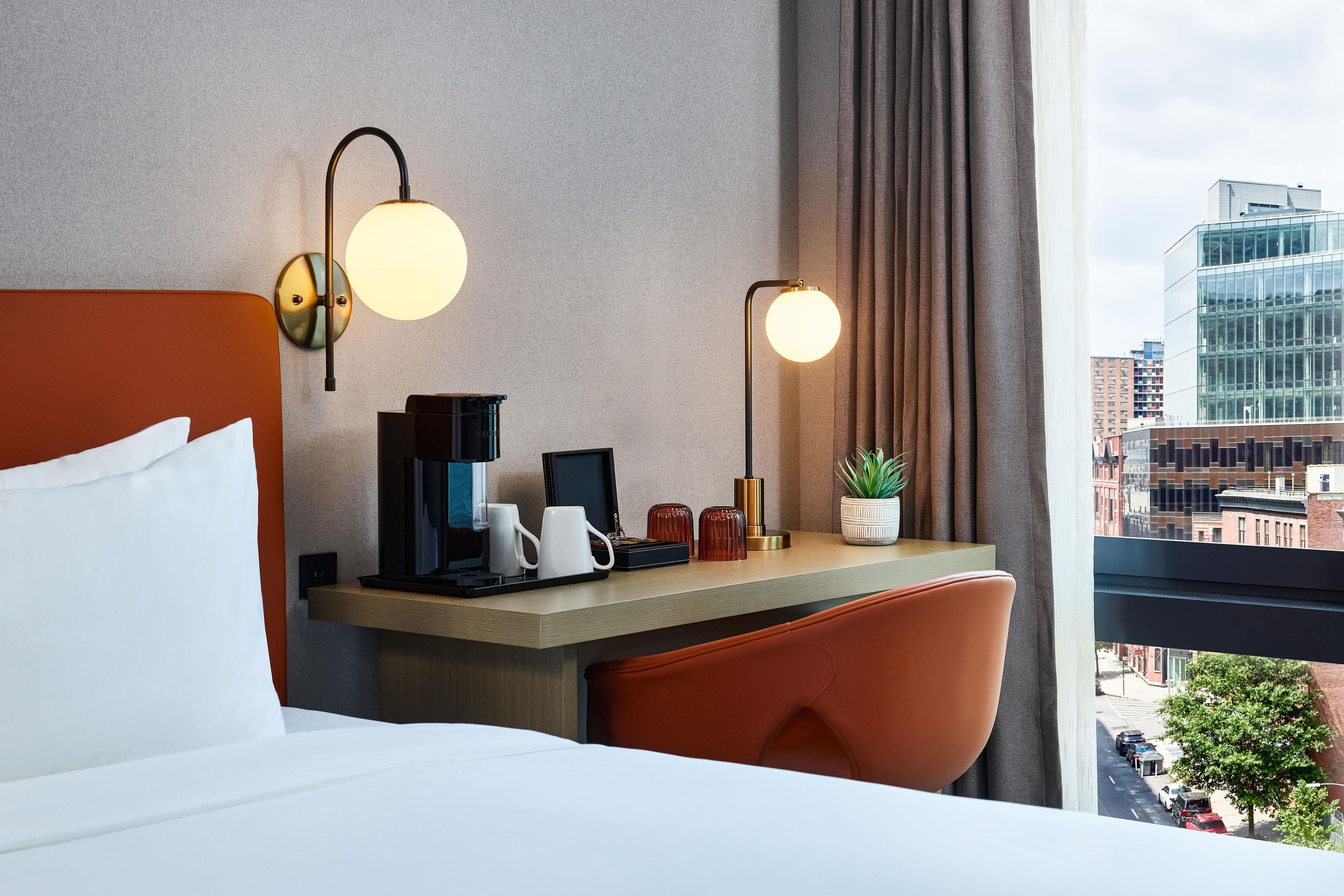 Quarto de hotel com cama, secretária com máquina de café, duas luminárias e vista para a cidade.
