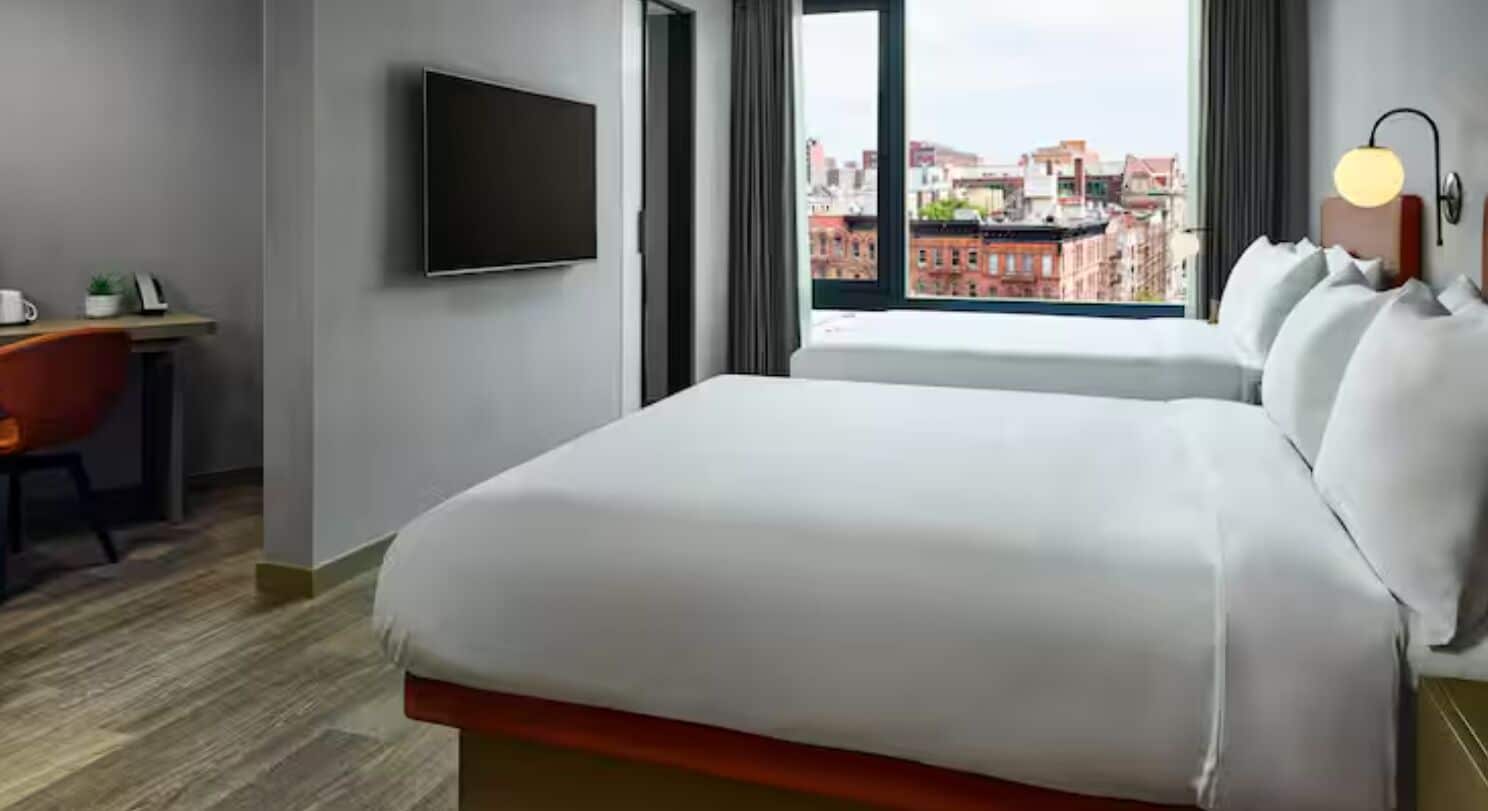 A hotel room with a bed, a desk, a television, and a window with a city view.