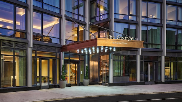 Entrada moderna d'un hotel amb façana de vidre, un rètol que diu "The George Manhattan", i una zona d'entrada coberta amb plantes en testos.