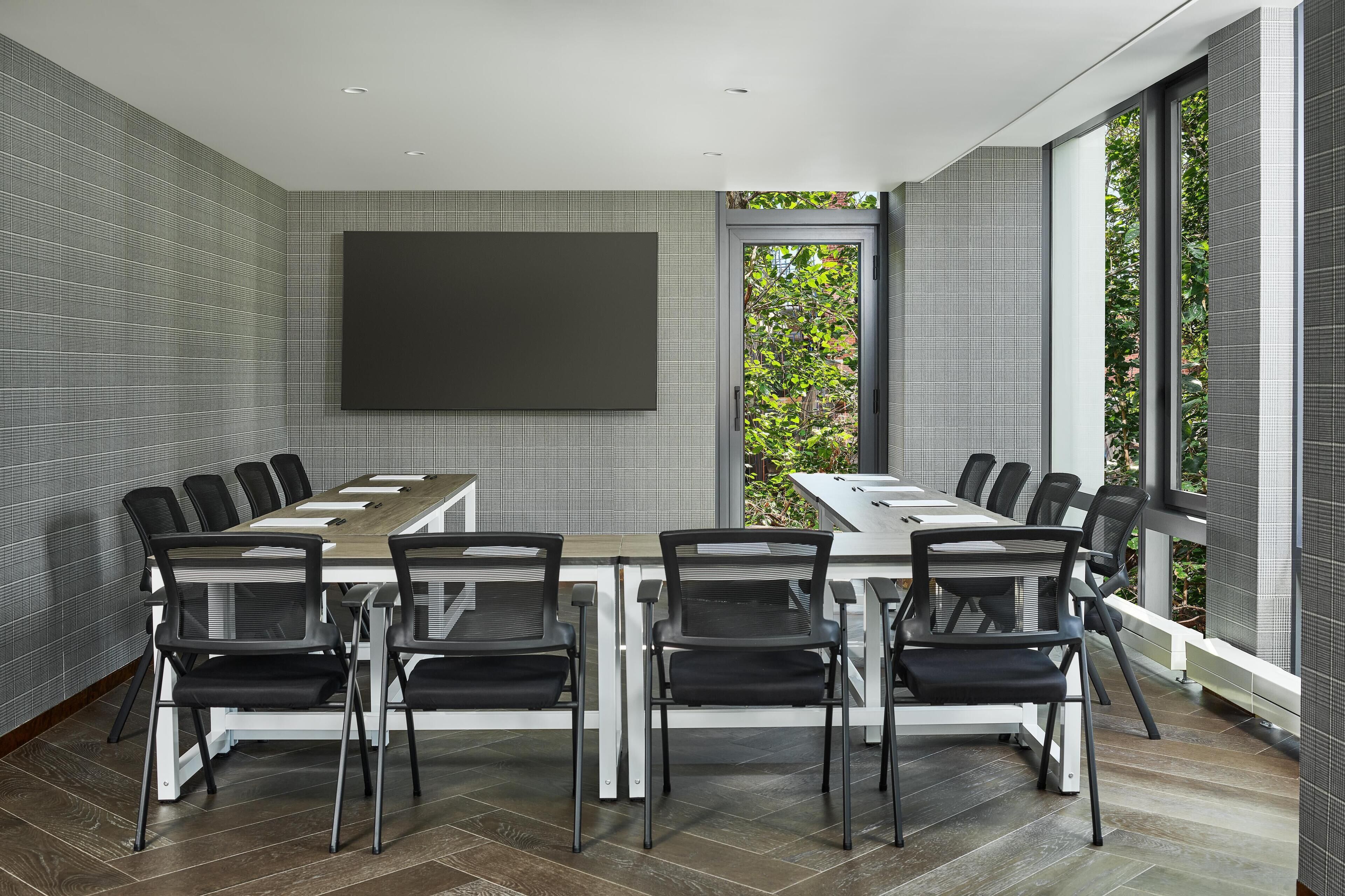 A modern meeting room with a long table, black chairs, a large screen, and large windows with a view of greenery.