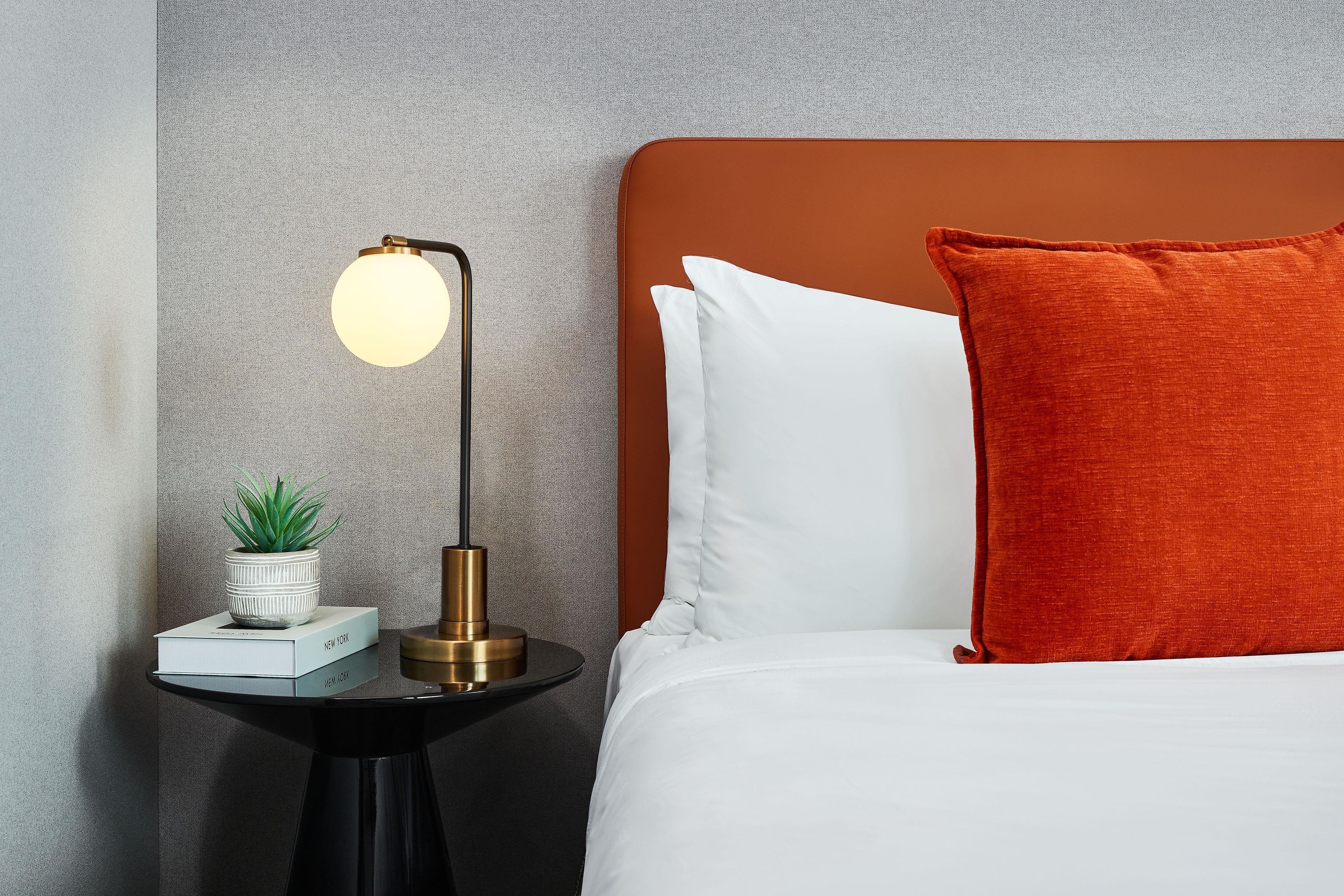 Un lit bien fait, agrémenté d’oreillers blancs et orange, une petite table de chevet avec une lampe, une plante en pot et un livre intitulé « NEW YORK ».