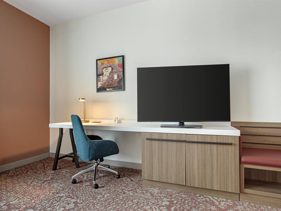A hotel room with a desk, chair, television, and a bench.