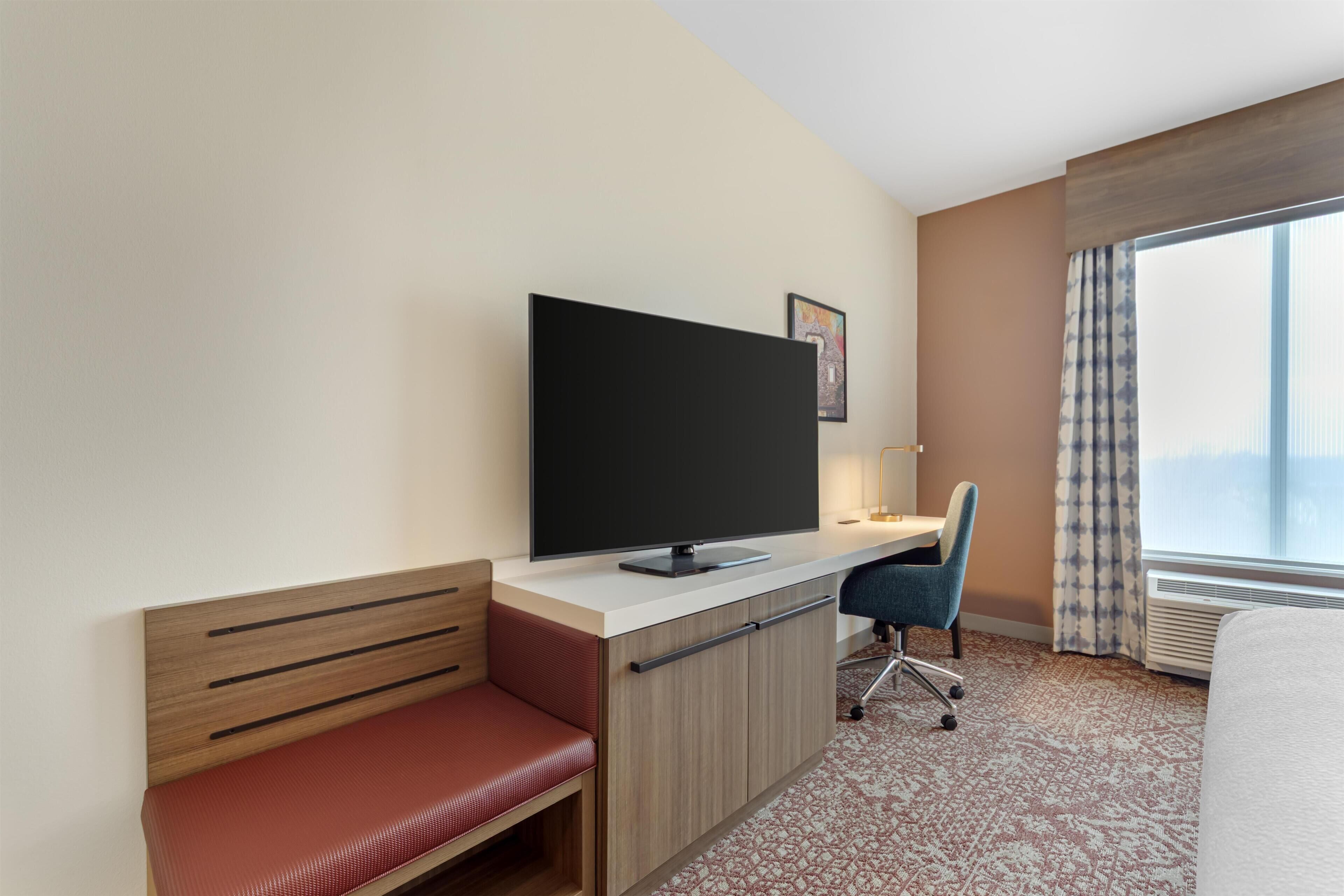 Ein Hotelzimmer mit einem Bett, einem Schreibtisch mit Flachbildfernseher, einer roten Bank, einem Sessel und einem großen Fenster mit Vorhängen.