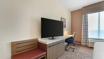 Ein Hotelzimmer mit einem Bett, einem Schreibtisch mit Flachbildfernseher, einer roten Bank, einem Sessel und einem großen Fenster mit Vorhängen.