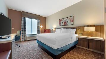 A hotel room with a bed, a desk, a chair, a television, a window with curtains, and a painting on the wall.