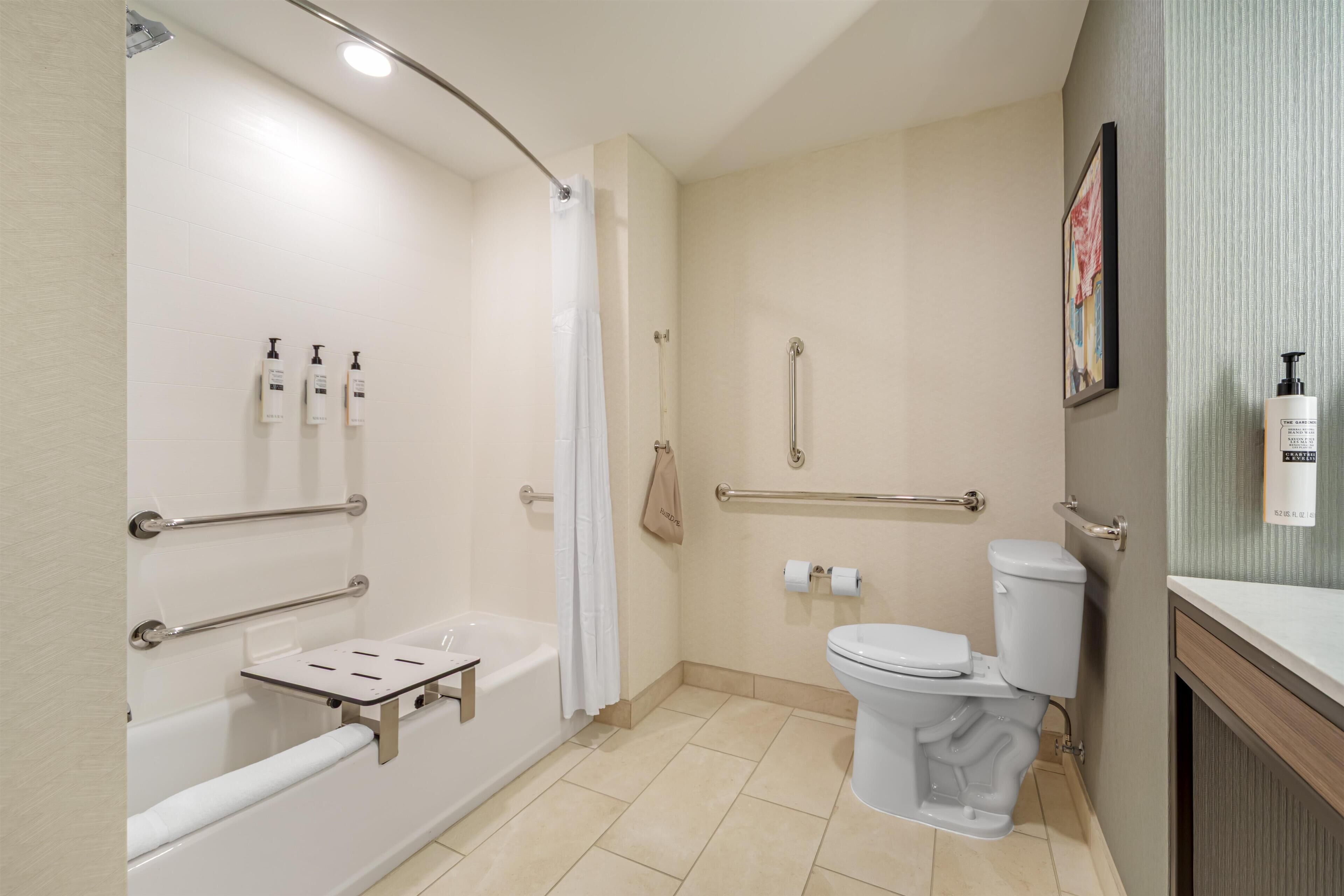 A bathroom with a bathtub, toilet, and shower. There are handrails, a towel rack, and a toilet paper holder.