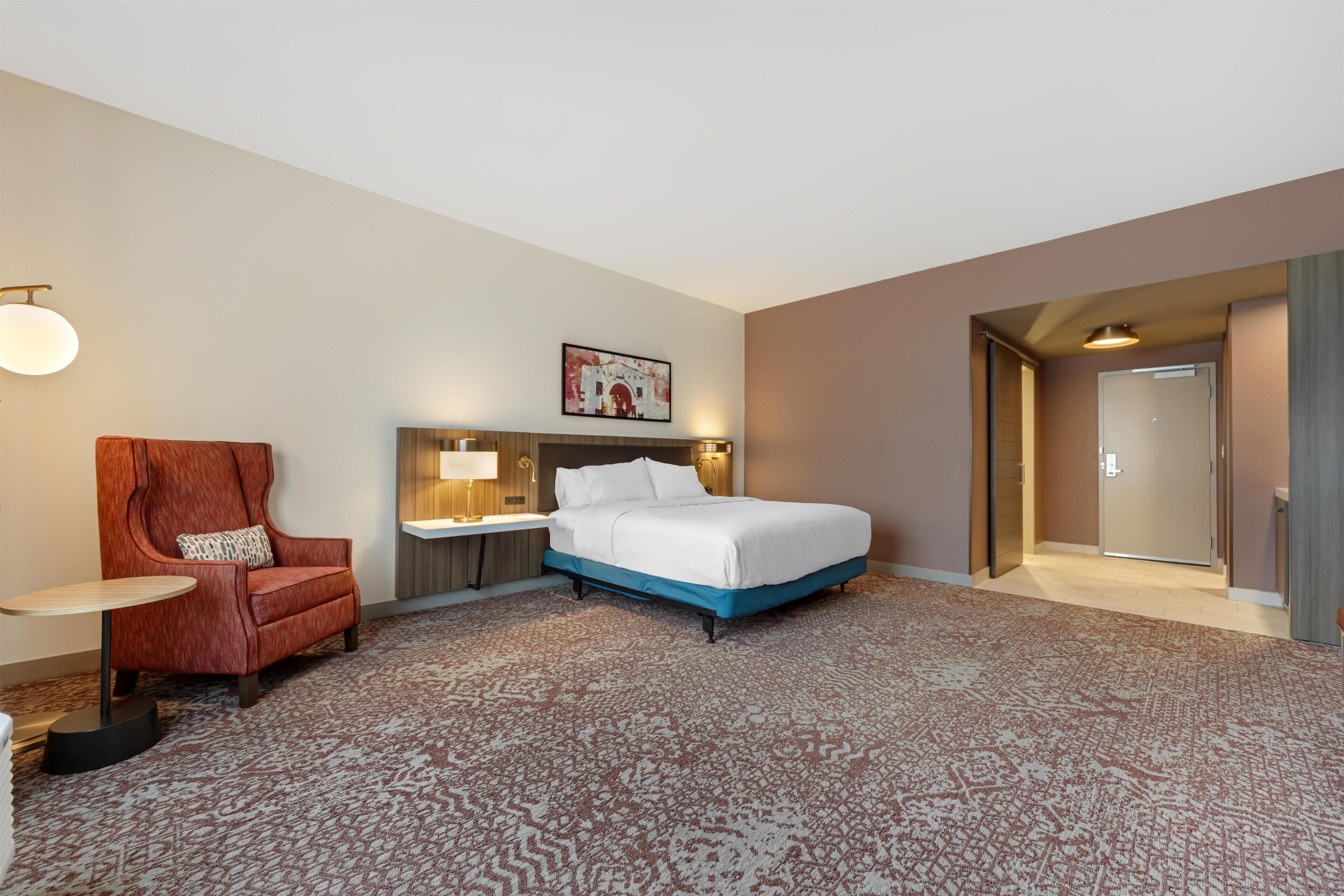 Una habitación de hotel con cama, una silla, una mesita de noche, una lámpara y una puerta.