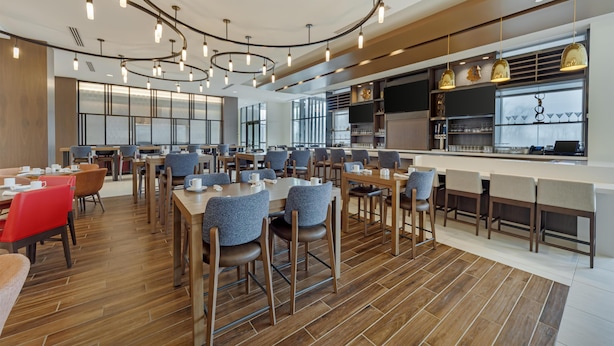 A modern dining area with wooden flooring, tables, and chairs, and a kitchen area with bar seating. - Hilton Garden Inn Media Philadelphia (Media)