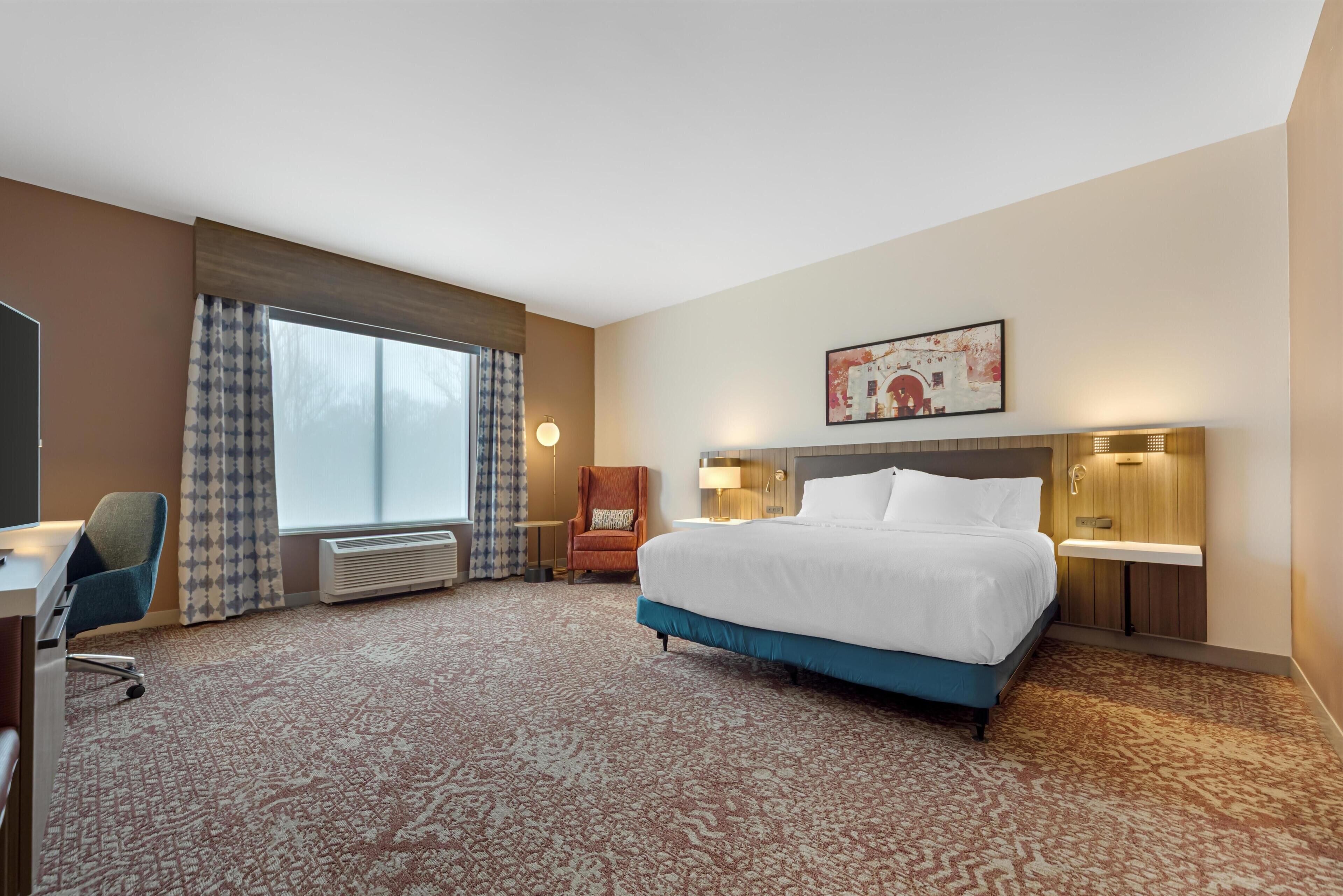 A hotel room with a large bed, a desk, a chair, a window with curtains, a wall-mounted artwork, and a wall-mounted light fixture.