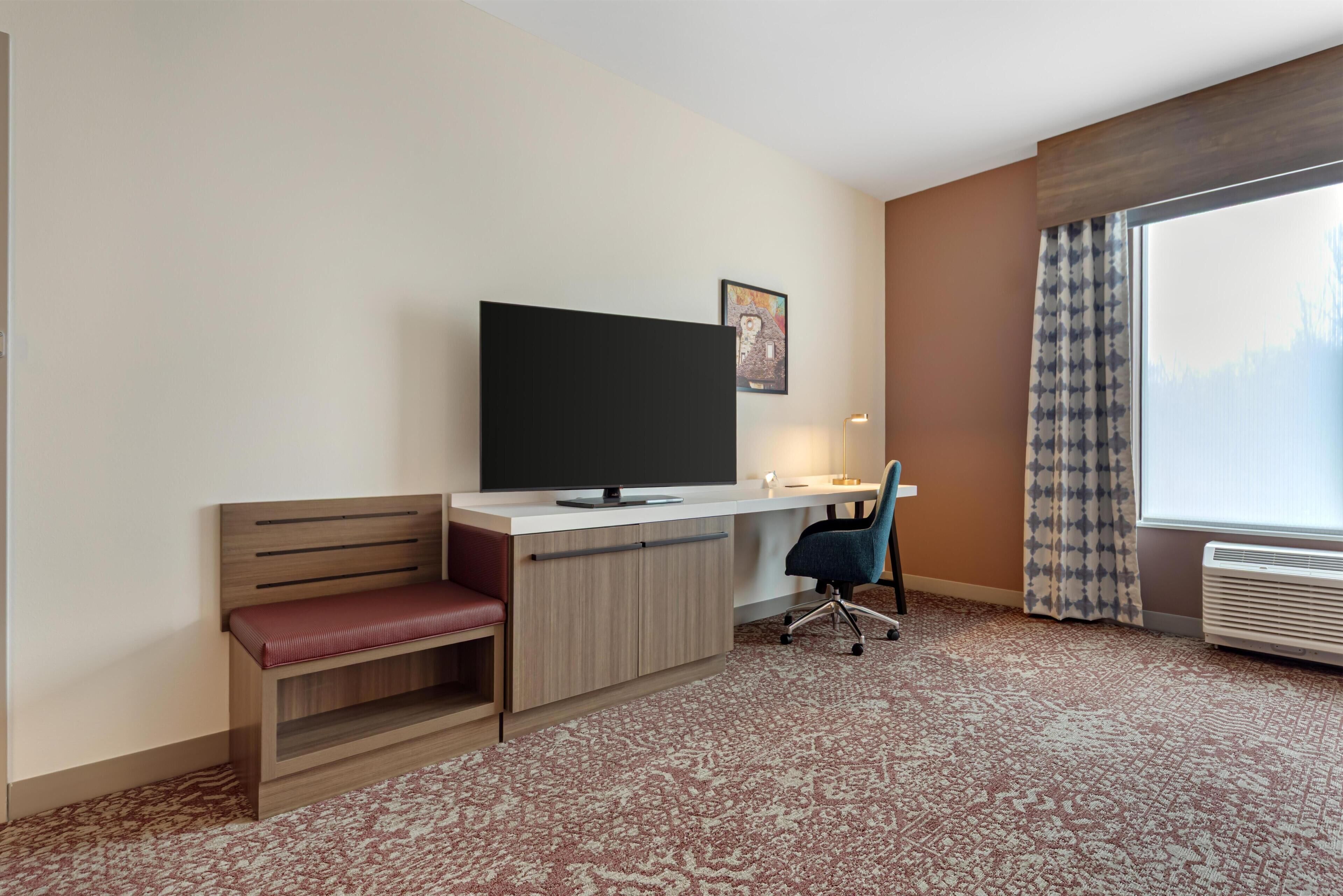 Quarto de hotel com escrivaninha, televisão, cadeira, banco e uma janela grande.