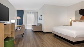 A hotel room with a bed, a desk with a chair, a television, and a green ottoman.