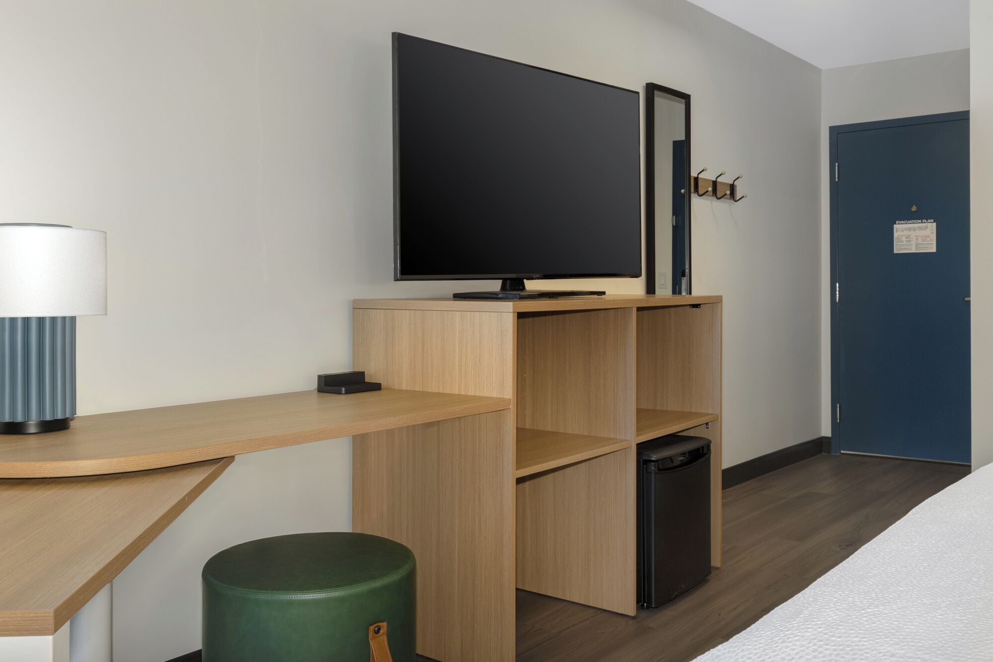 Une chambre d’hôtel moderne équipée d’une télévision à écran plat, d’un bureau avec une lampe, d’un pouf vert et d’une porte bleue.