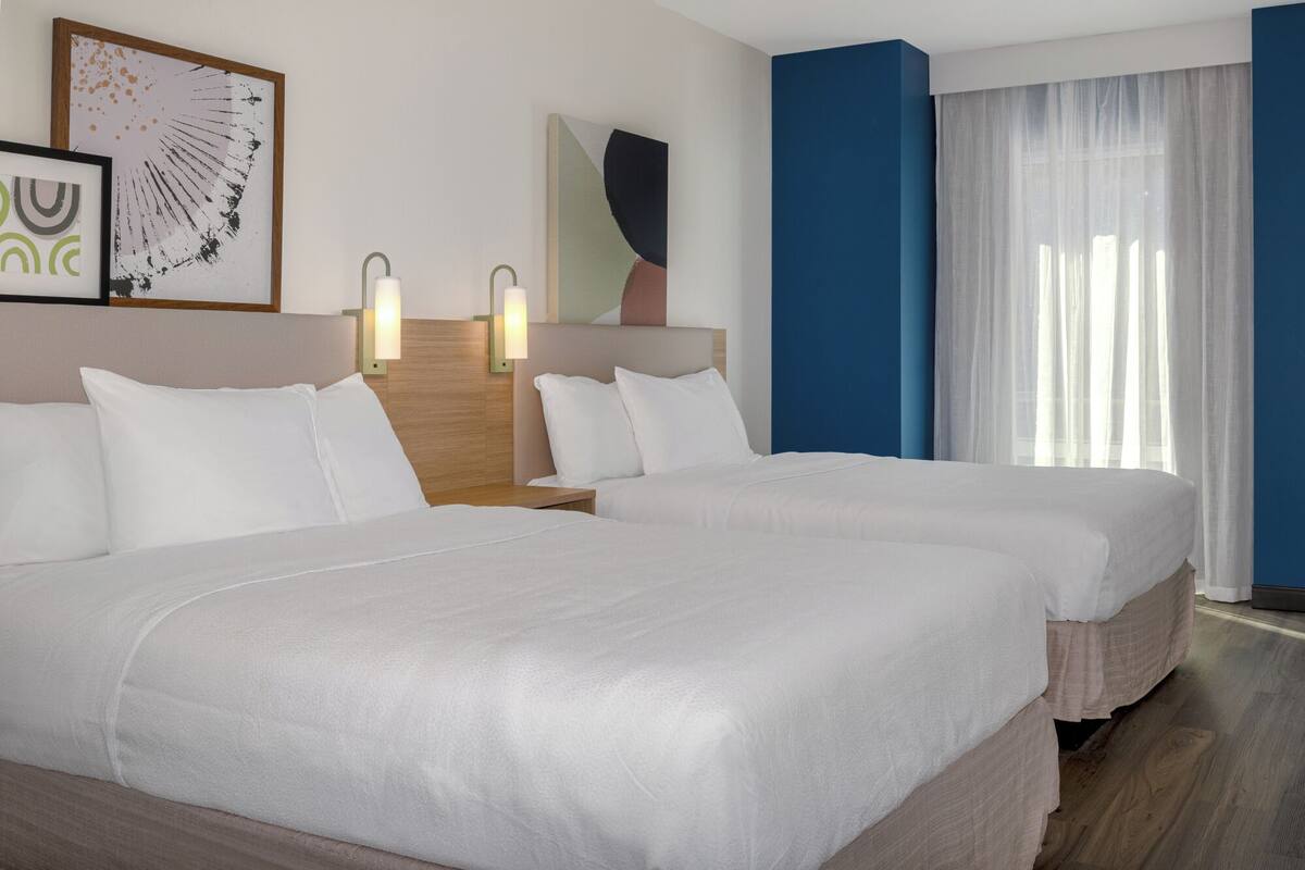 Ein Hotelzimmer mit zwei Betten, einem Holzkopfstück, einem großen Fenster mit durchsichtigen Vorhängen und Wandbildern.