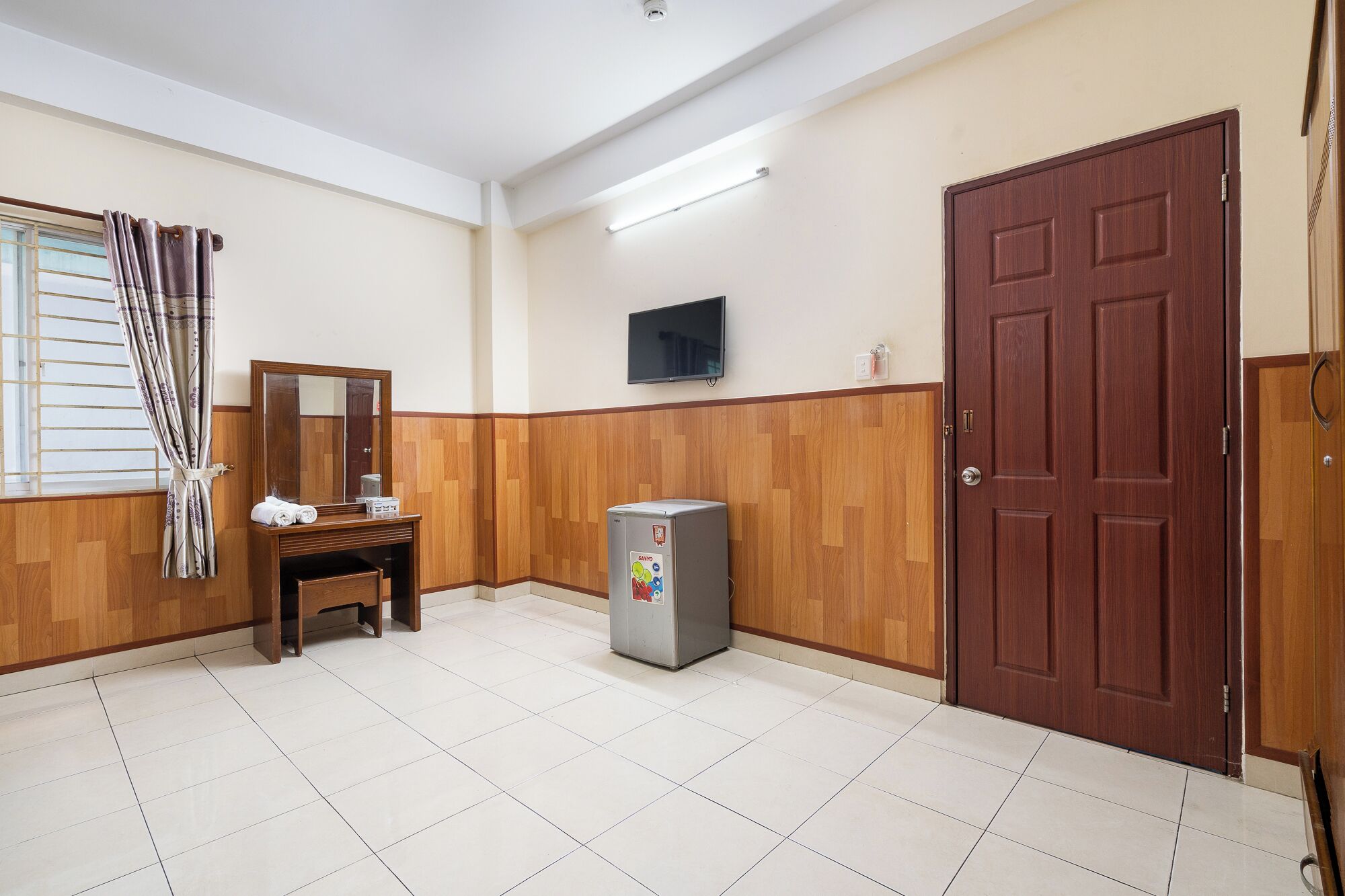 A hotel room with a wooden panel wall, a large mirror, a small table with a chair, a refrigerator, a wooden door, and a window with curtains.