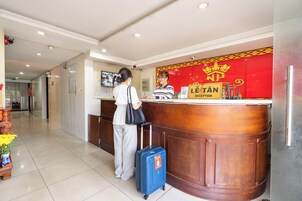 Uma área de recepção de hotel com uma mesa de atendimento em madeira, uma placa vermelha e dourada indicando a recepção, e uma mulher em pé no balcão com uma mala.