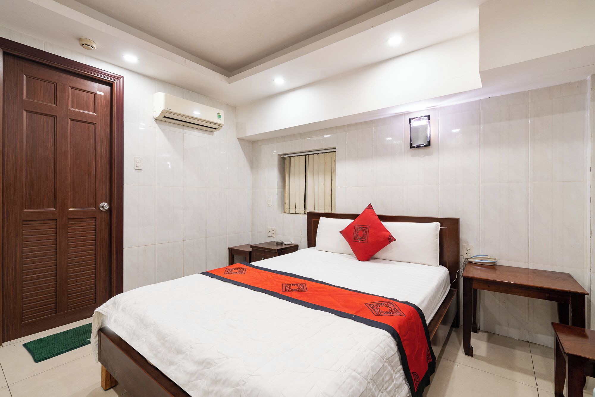 A hotel room with a wooden door, a bed with a white and red bedspread, a nightstand, a bedside table, and a wall-mounted air conditioner.