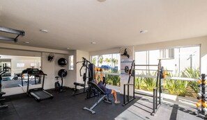 Un gimnasio con varios equipos de ejercicio, un espejo grande y vistas a palmeras fuera.