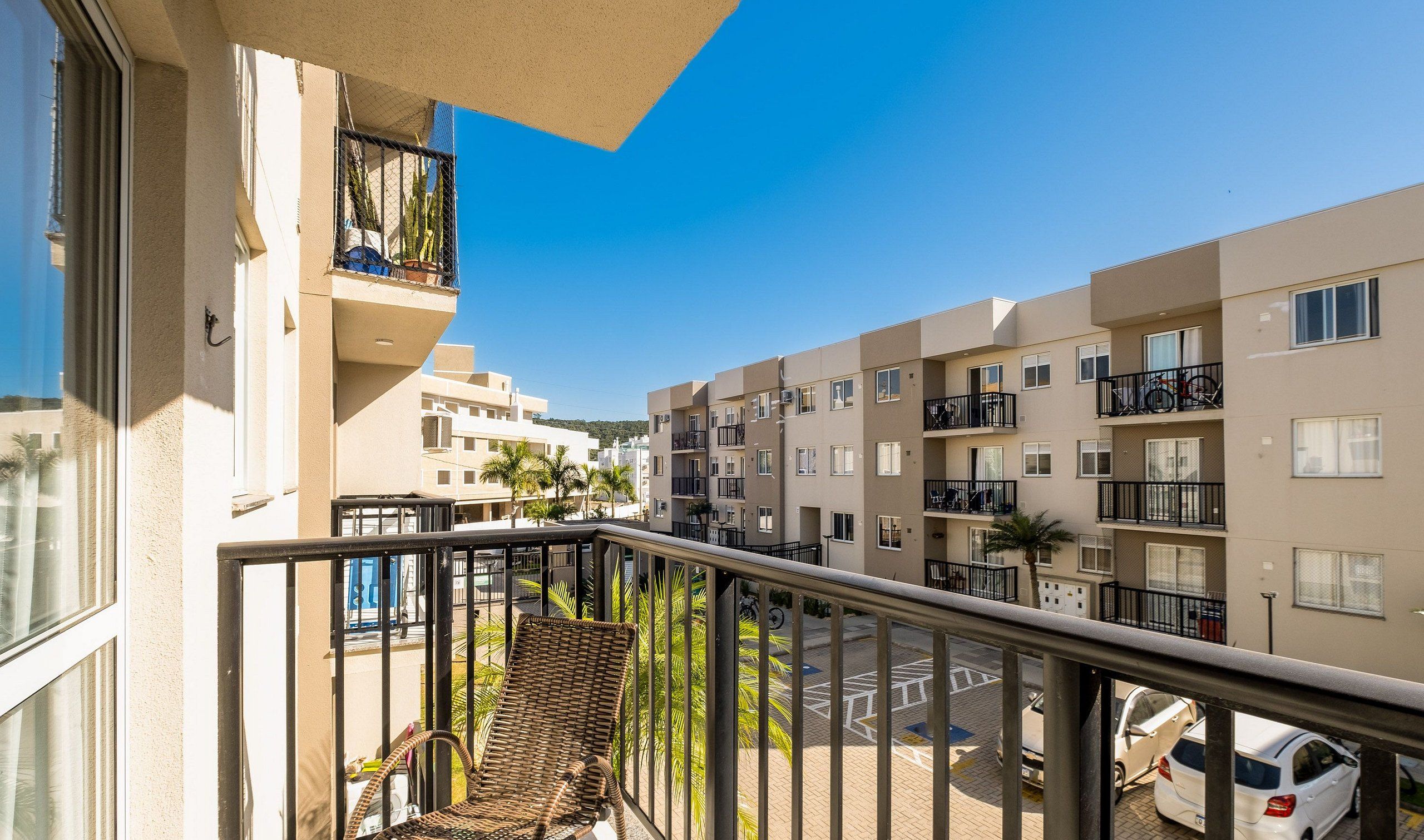 Un balcón con una silla de mimbre, una planta en maceta y vistas a una zona residencial con varios edificios de apartamentos.