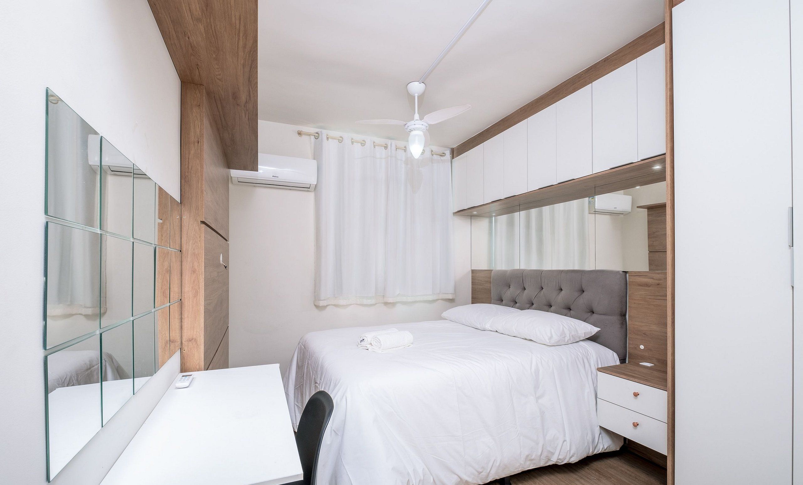 A bedroom with a bed, a desk, a mirror, and a ceiling fan.