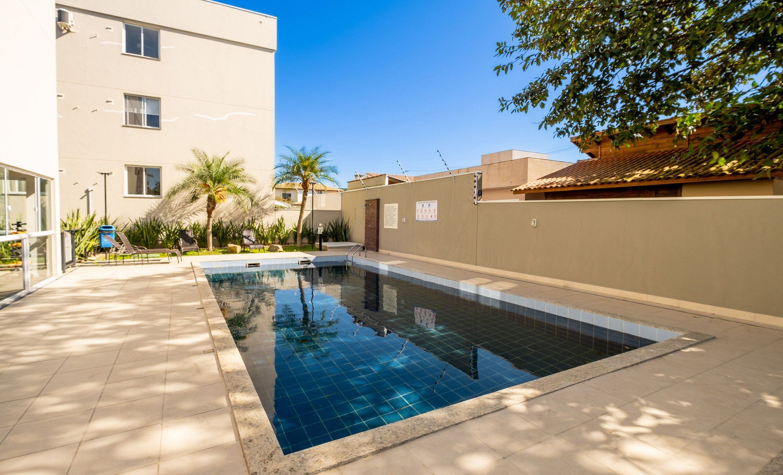 Une piscine dont les bords sont recouverts de carreaux, entourée d’un bâtiment de couleur beige et de palmiers.