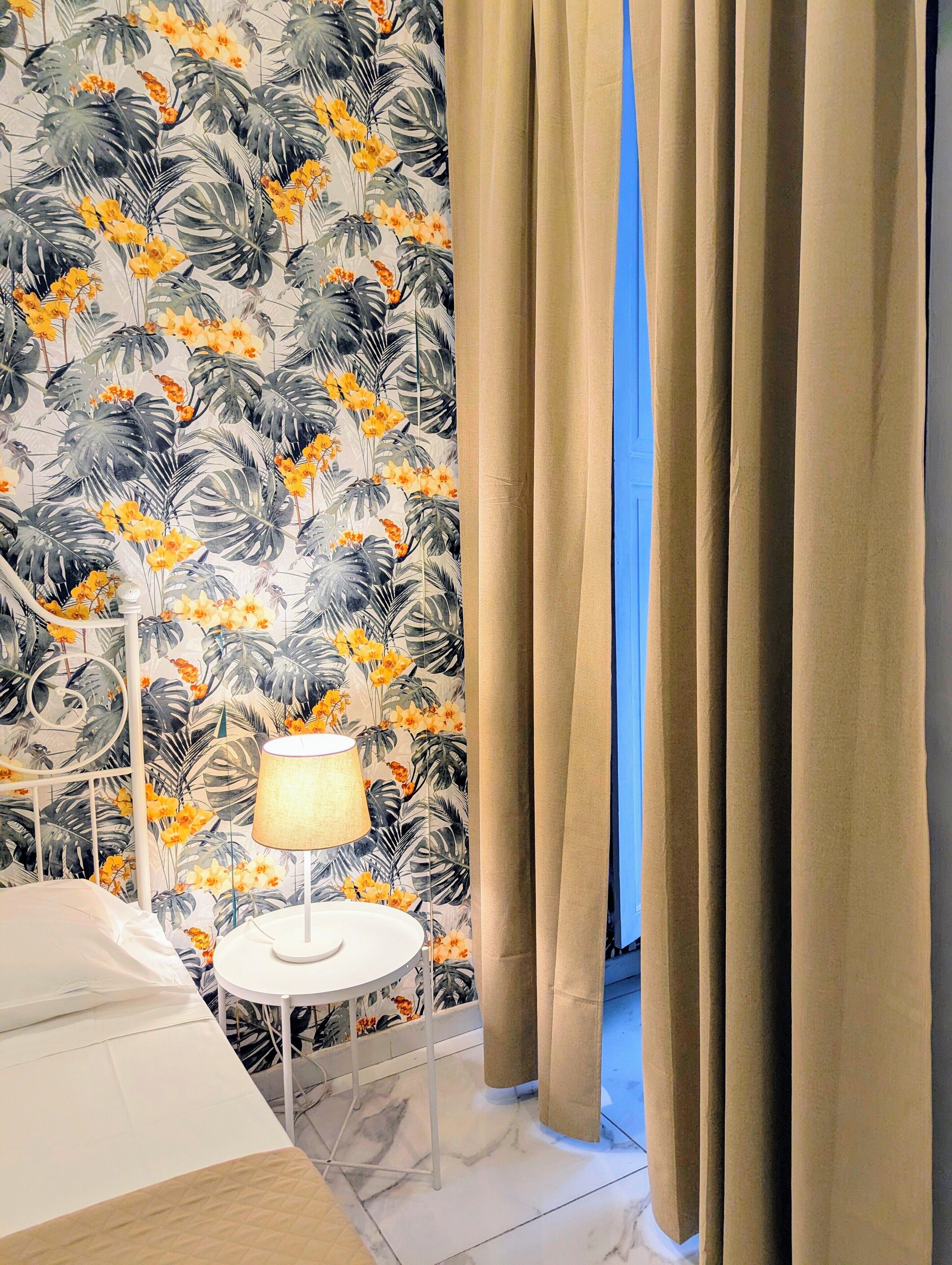 A hotel room with a bed, a small round table with a lamp, a window with beige curtains, and a wall with a tropical leaf and flower wallpaper.
