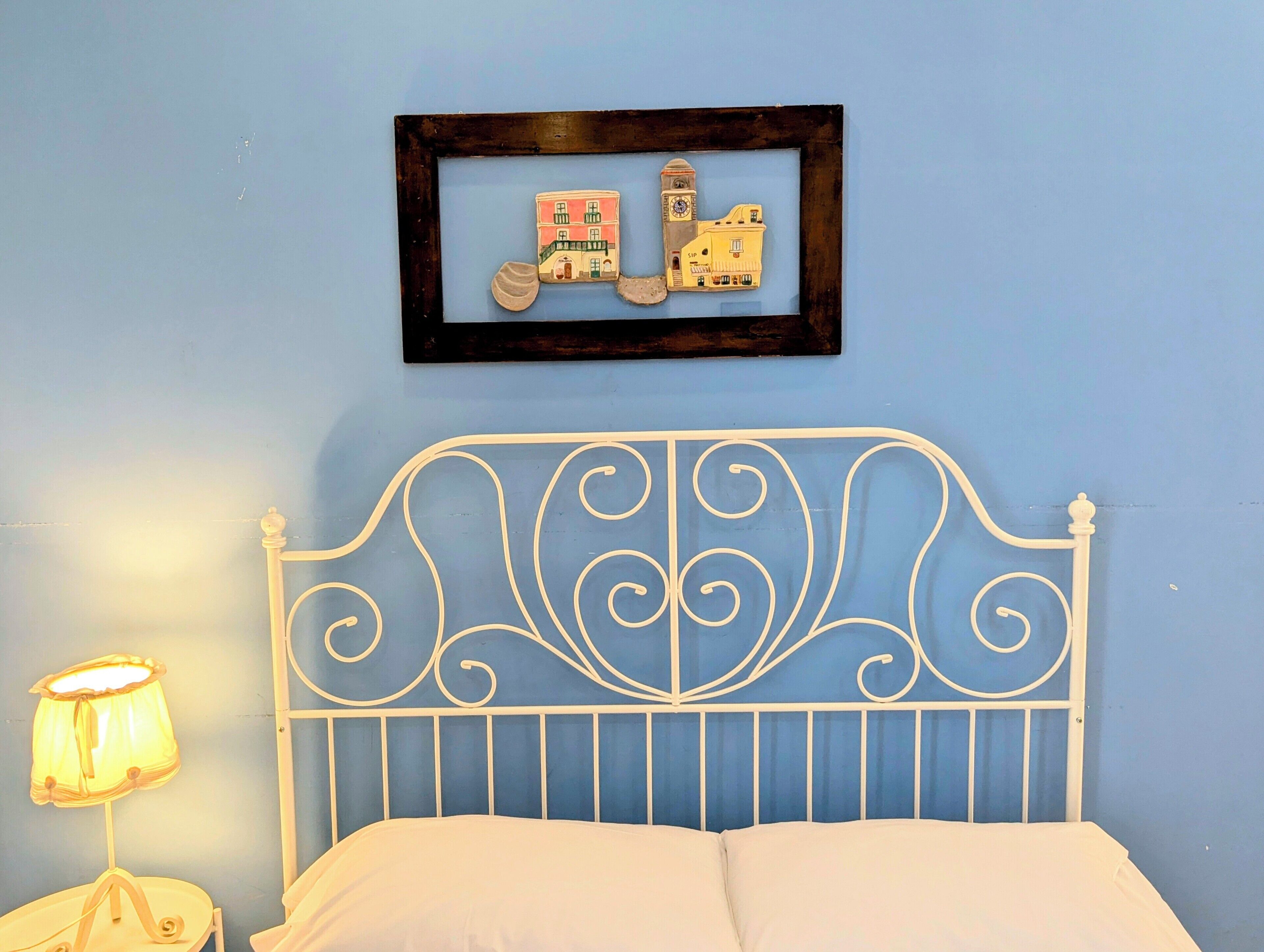 Un dormitorio con pared azul, estructura de cama metálica blanca, cubrecama blanco, un cuadro enmarcado con edificios y una lámpara de noche.