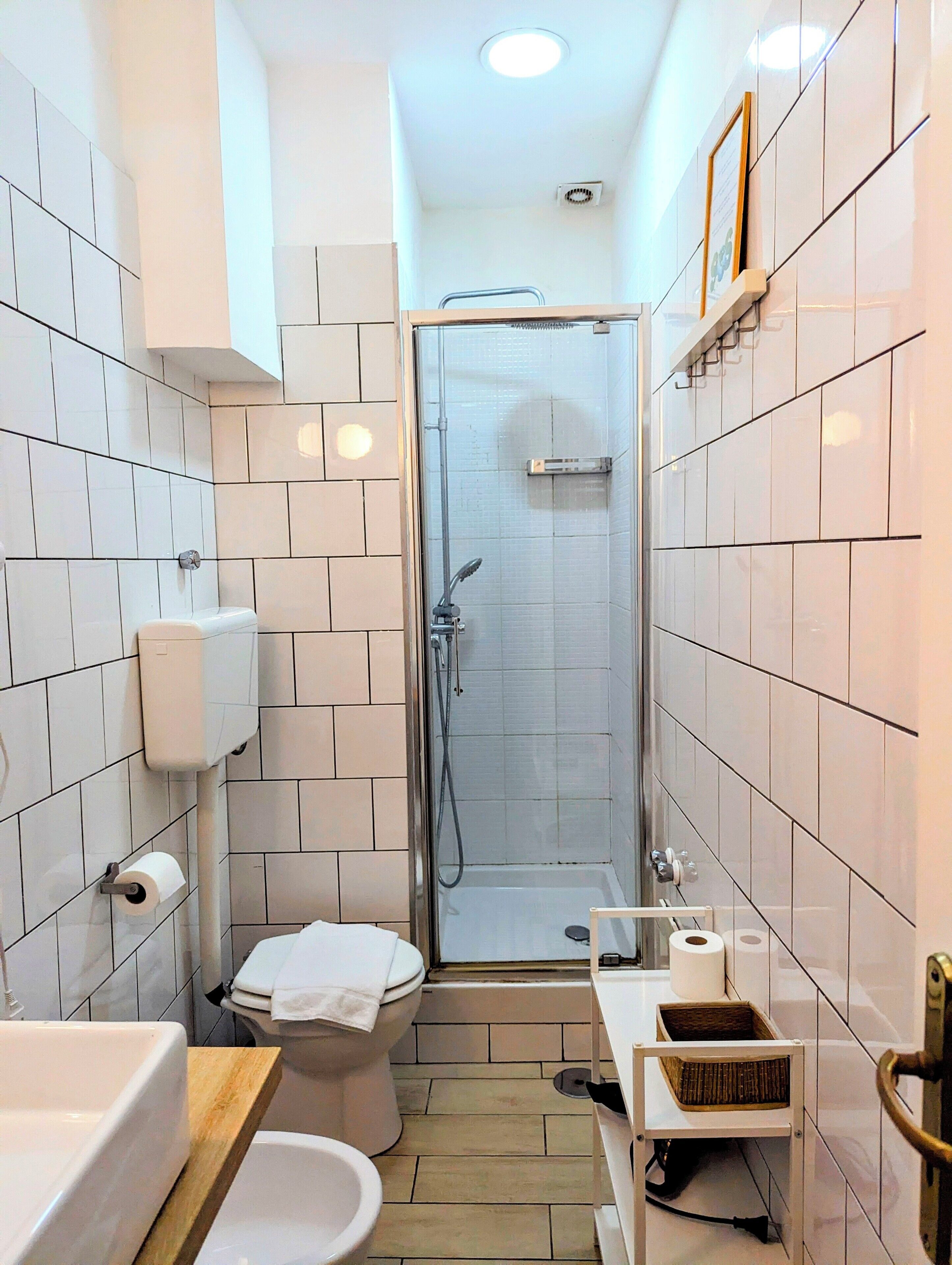 A modern bathroom with white tiles, a glass-enclosed shower, a toilet, a sink, and a small storage basket.