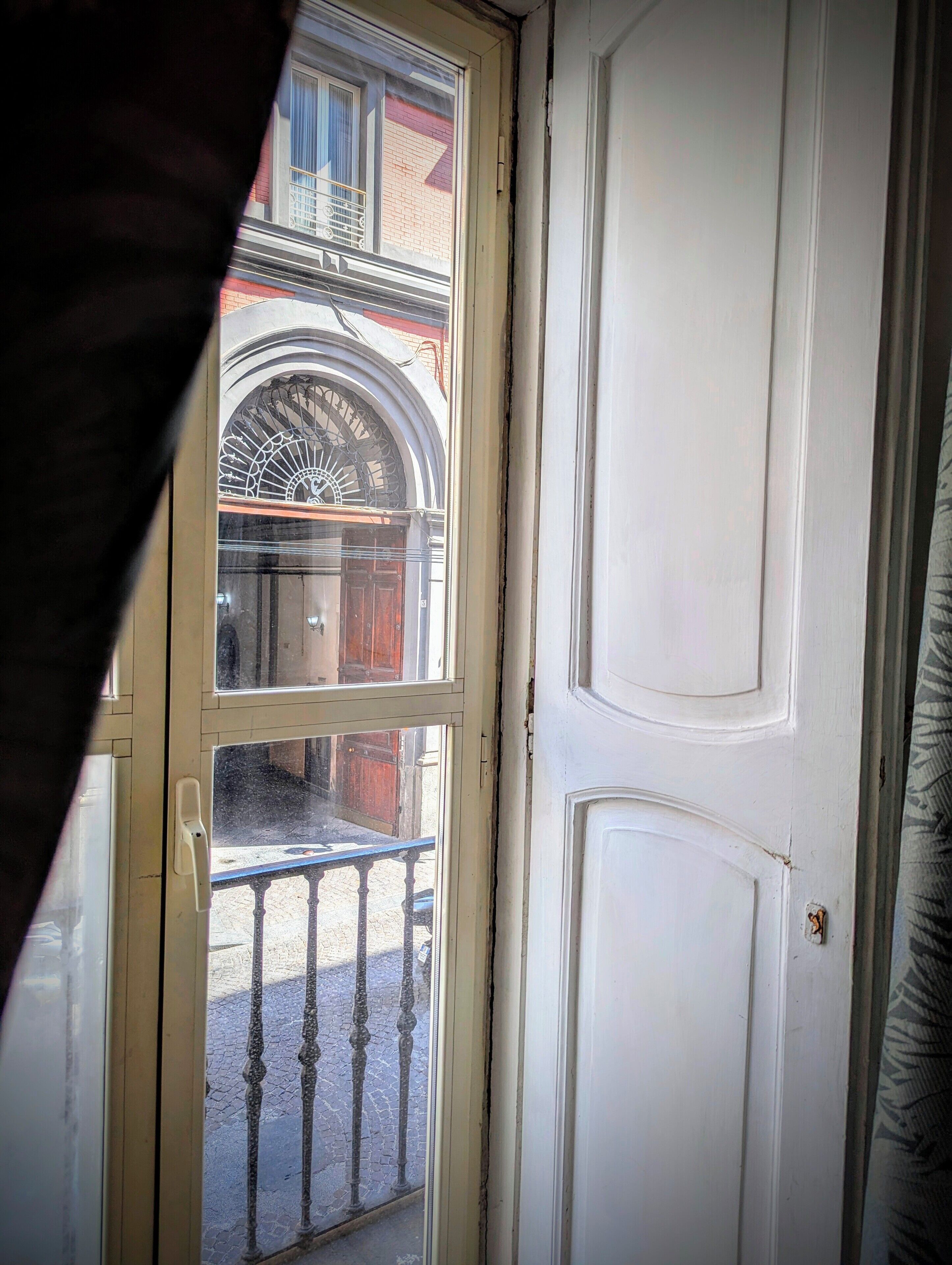 Eine Tür mit Glasfenster, hinter der sich ein Balkon mit schmiedeeisernem Geländer und ein Backsteinhaus erkennen lässt.